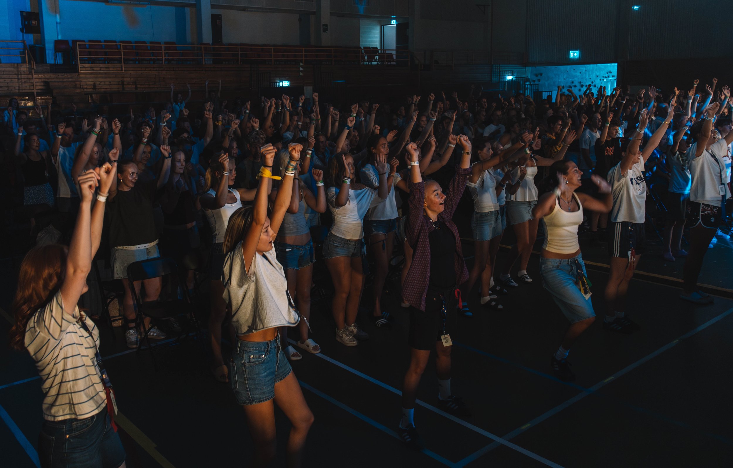 En stor gruppe mennesker i en innendørs arena, som deltar i en energisk aktivitet eller konsert, med armene hevet og entusiastiske.