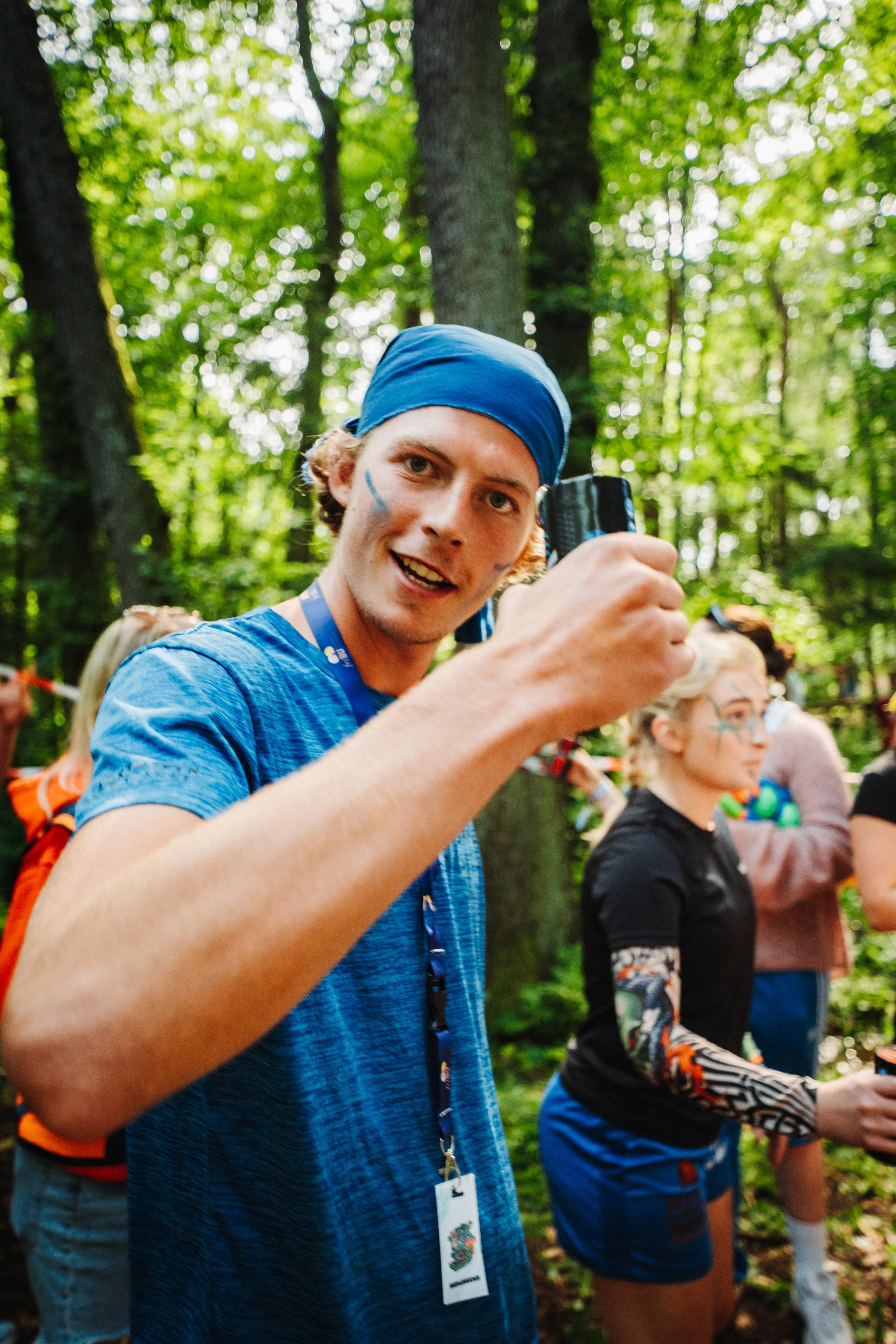 En gruppe mennesker i skogen iført fargerike klær og ansiktsmaling, med fokus på en mann i blå t-skjorte og blå hodebånd som smiler.