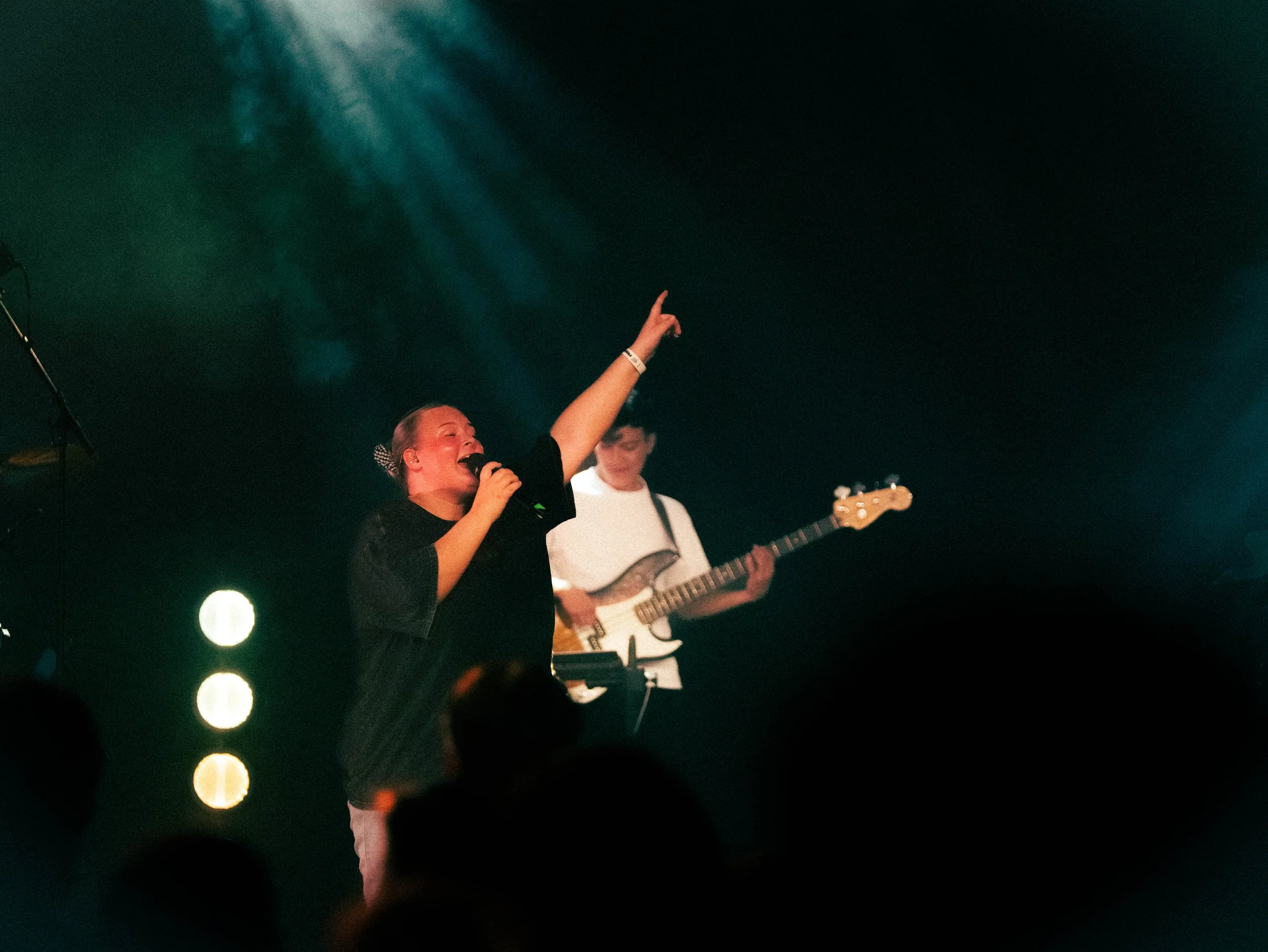 En jente synger inn i mikrofon på scenen, med en gitarist i bakgrunnen, under konsert med scenelys og mørk bakgrunn.