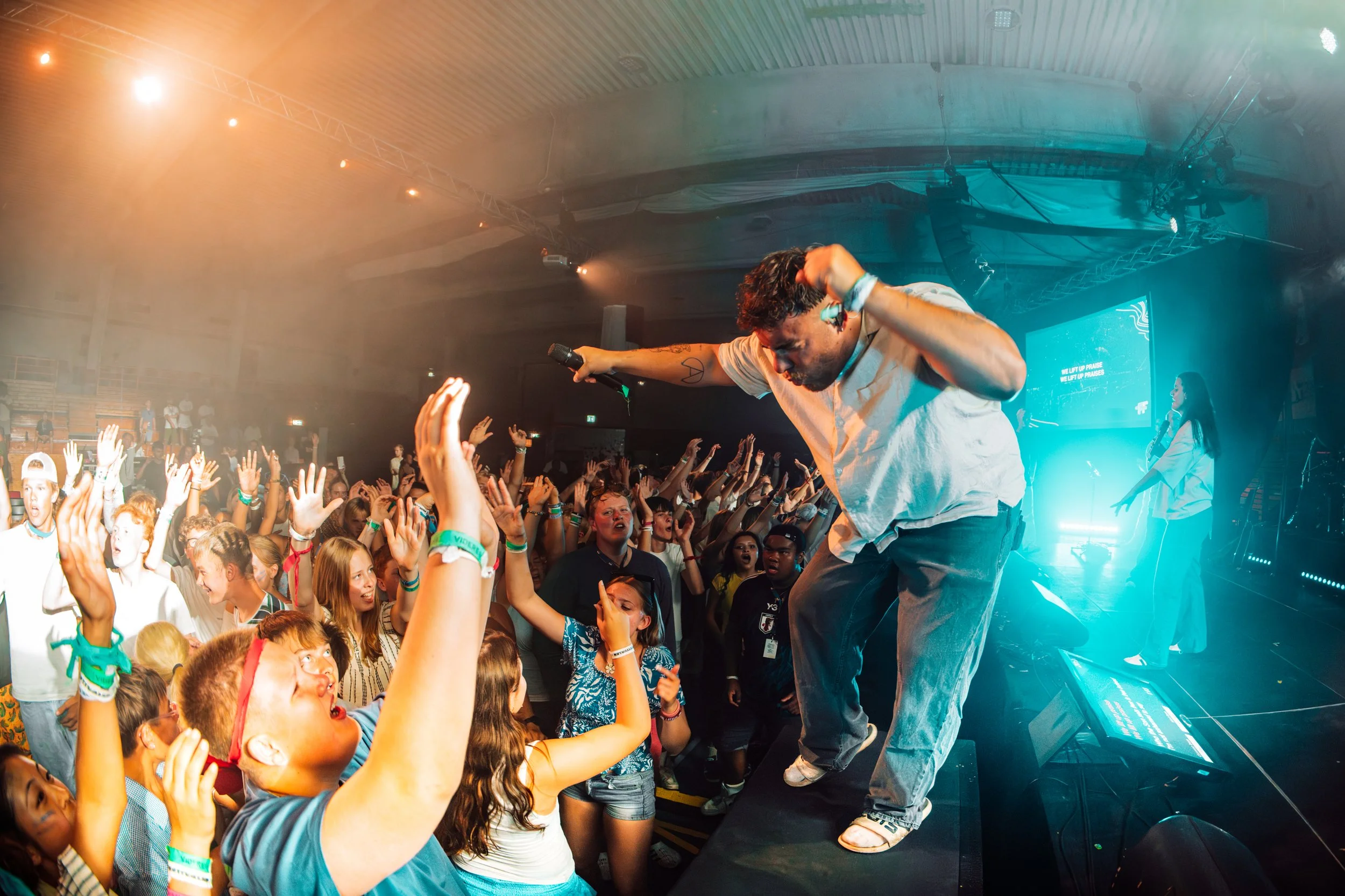 En mann på scenen som opptrer foran en engasjert publikum, i et innendørs konserthus med lyseffekter.