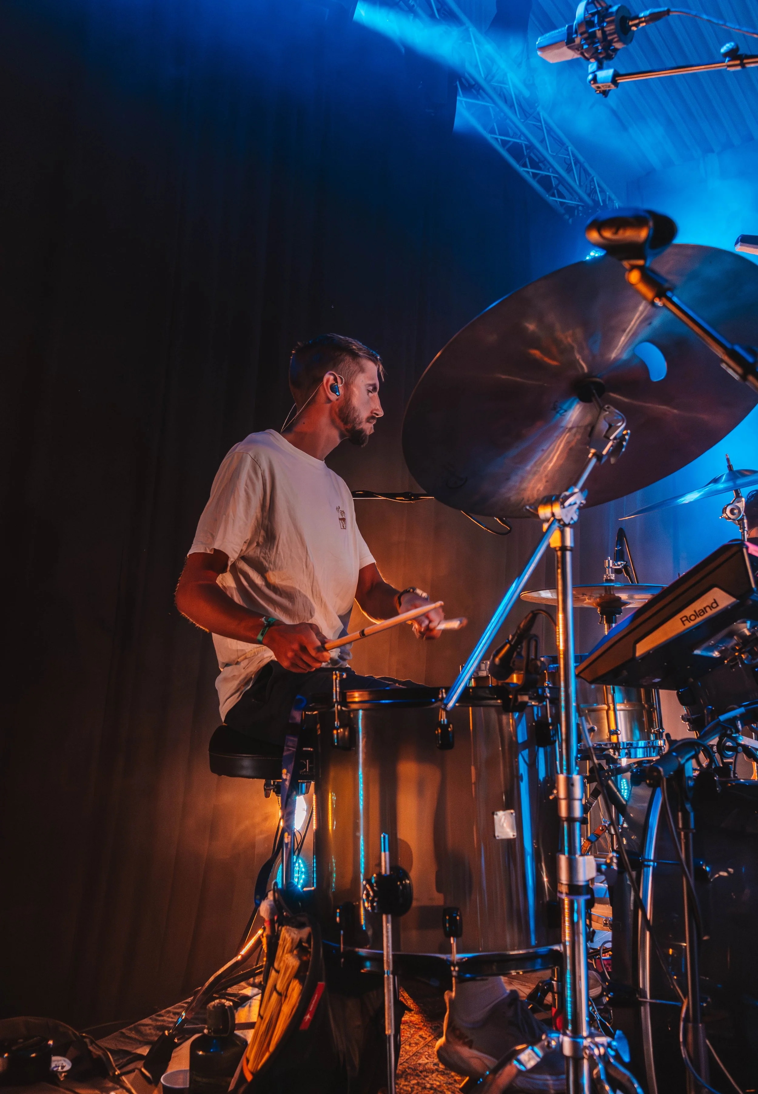 En mann som spiller trommer på scenen under en konsert, omgitt av musikkutstyr og blå belysning.