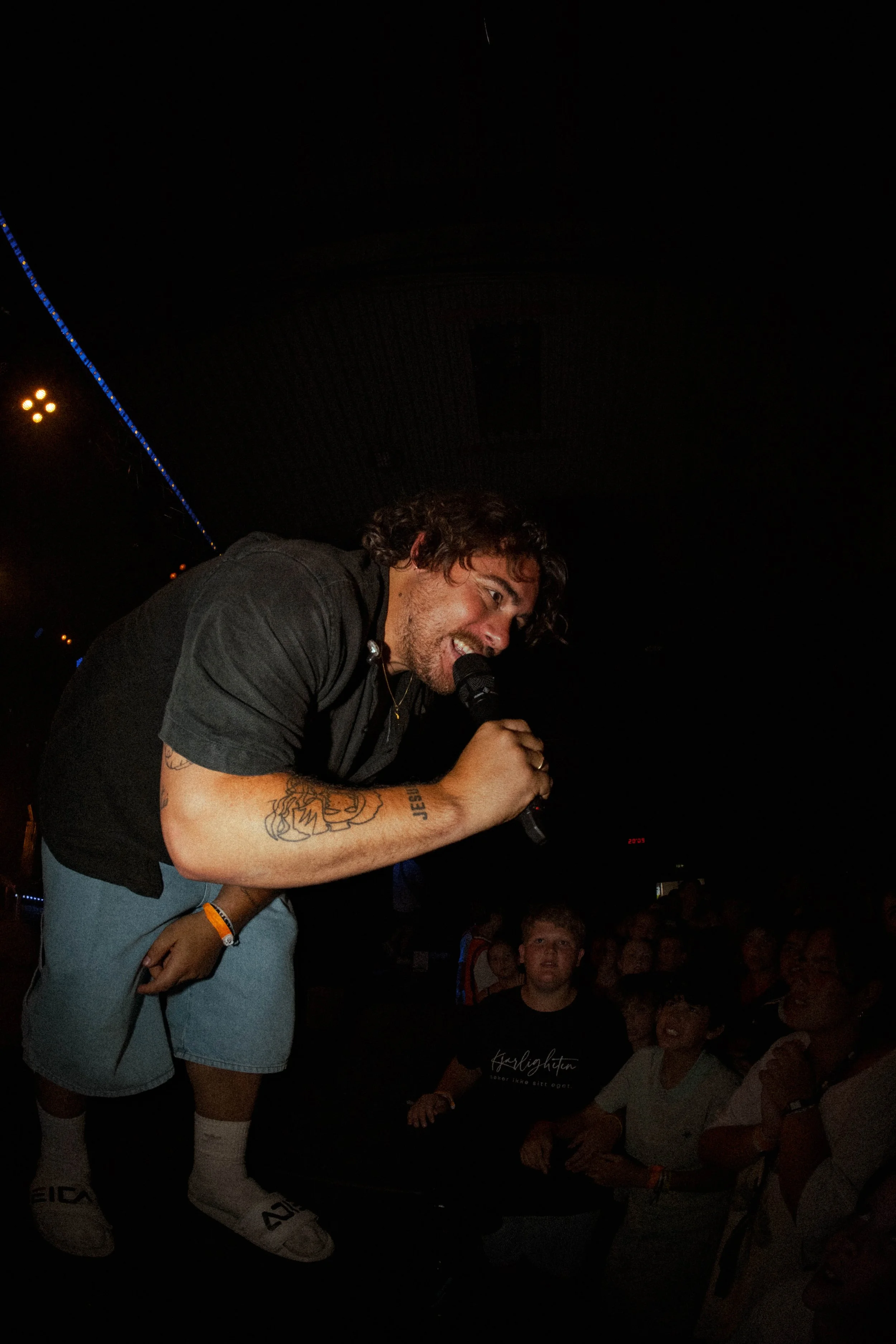 En mann med bølget hår holder en mikrofon og synger til et publikum på en mørk scene.