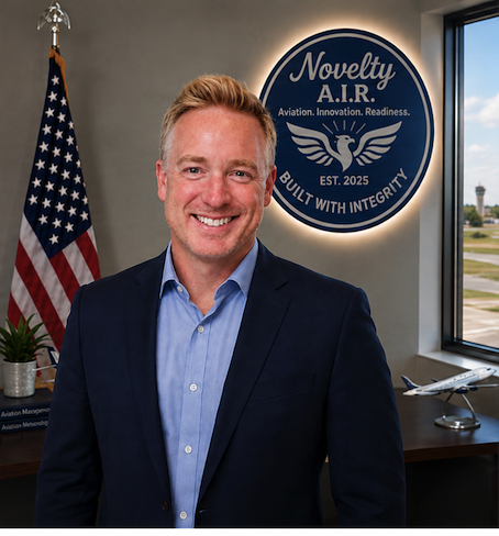 A smiling man in a navy suit and light blue shirt standing in an office with an American flag and a round sign that reads 'Novelty A.I.R. Aviation. Innovation. Readiness. Built with integrity. Est. 2025'. An airplane model is on a desk next to a window.
