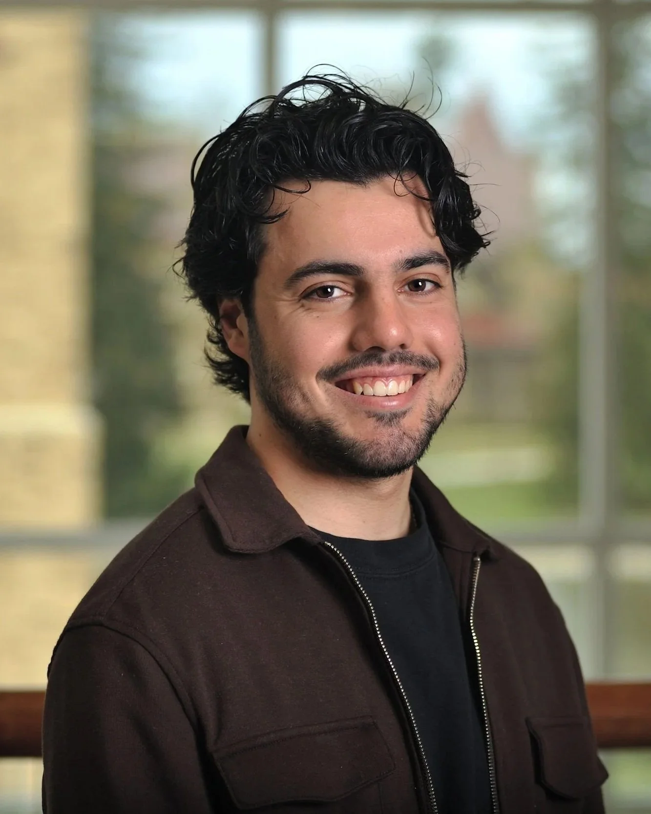 A smiling man with dark, wavy hair and a beard, wearing a dark brown jacket over a black shirt, standing indoors with a window and blurred outdoor scenery in the background.