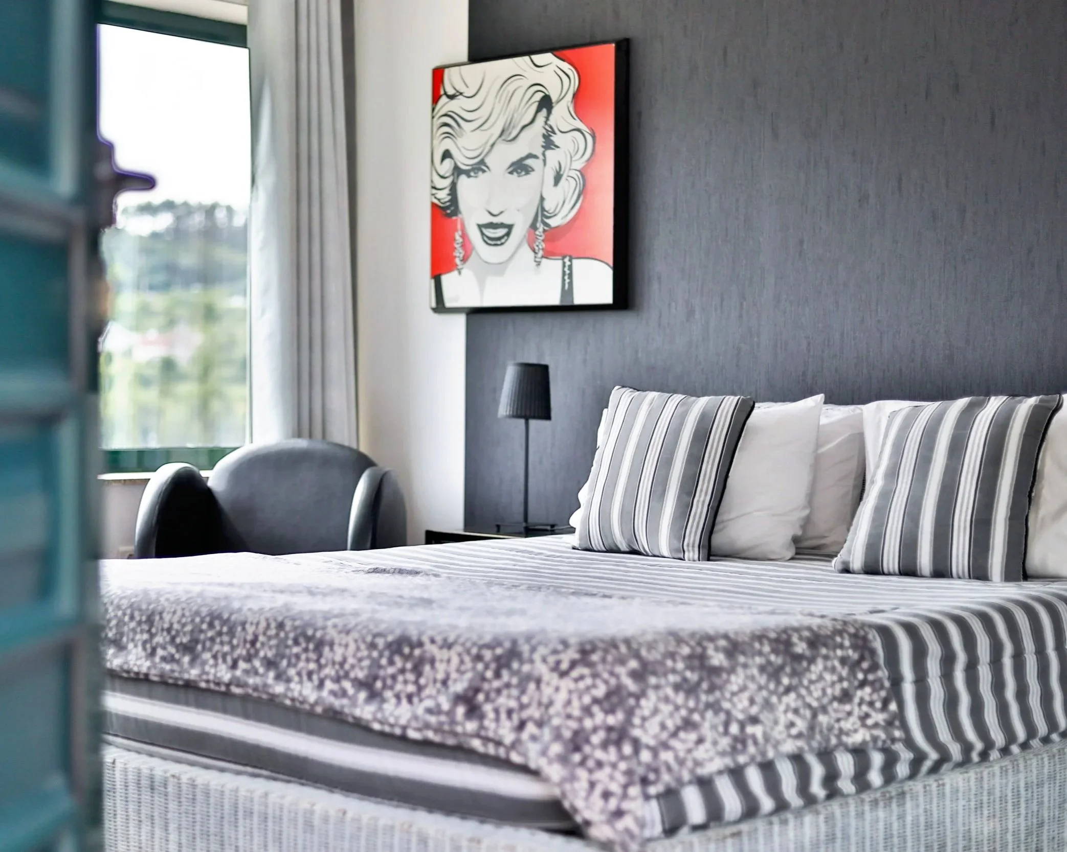 Quarto de hotel com cama de casal, almofadas listradas, quadro de arte de uma mulher famosa na parede, cadeira de leitura ao lado da janela com cortinas brancas, luminária de mesa ao lado da cama.