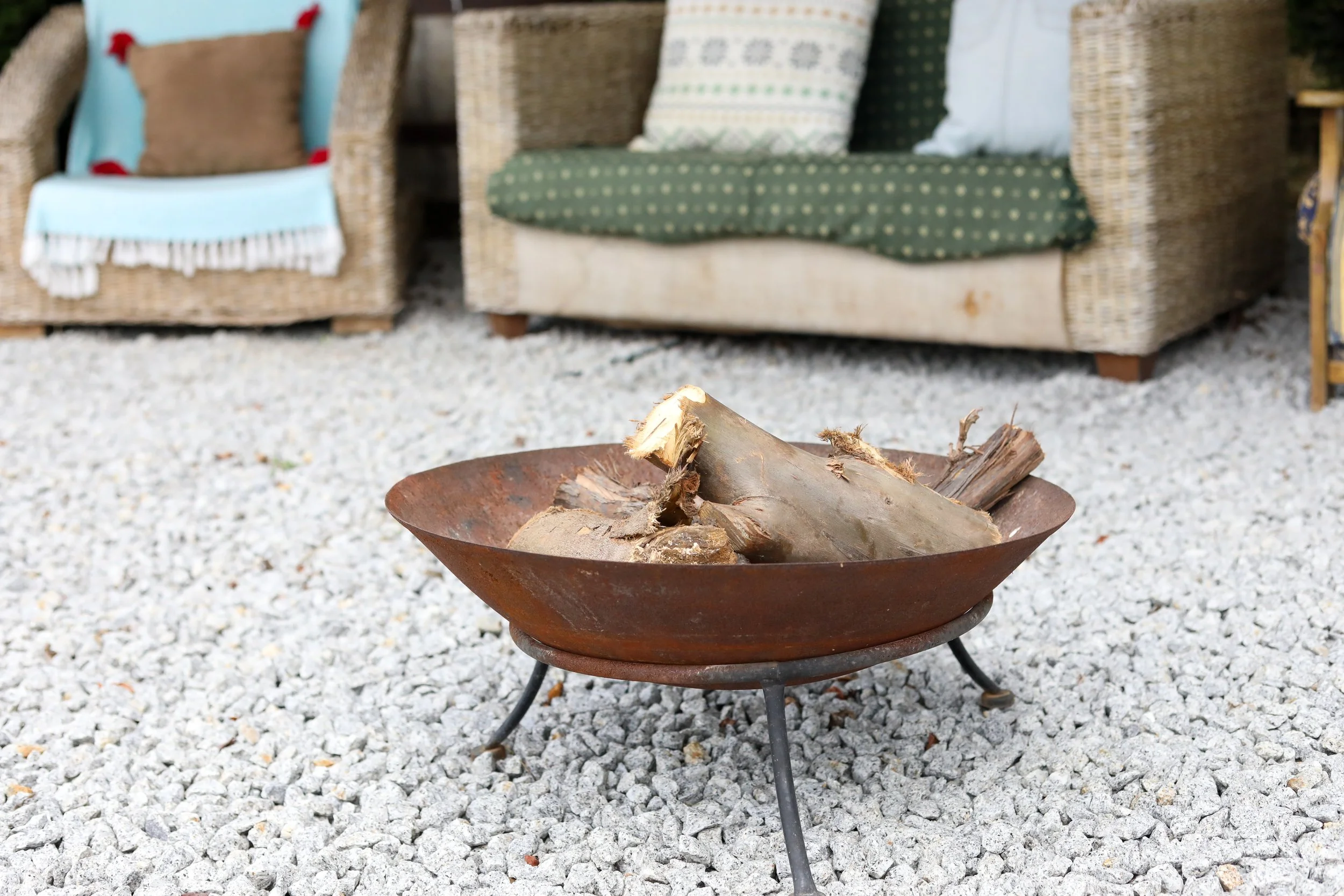Braseiro com carvão e pedaços de madeira, cercado por piso de pedra branca, com cadeiras de vime com almofadas ao fundo.
