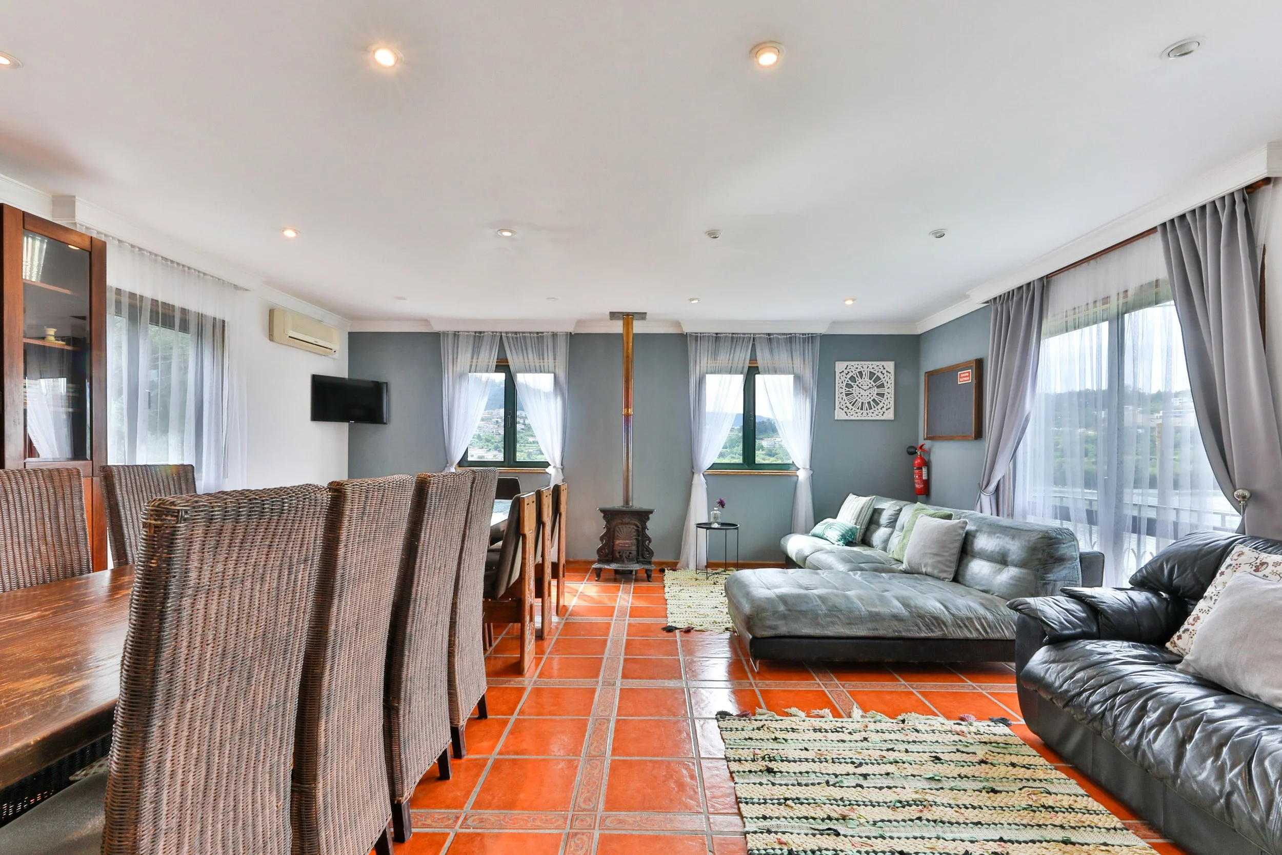 Sala de estar com piso de cerâmica vermelha, sofá cinza, sofá preto, janelas com cortinas brancas, mesa de centro, televisão na parede e quadro com detalhes decorativos.