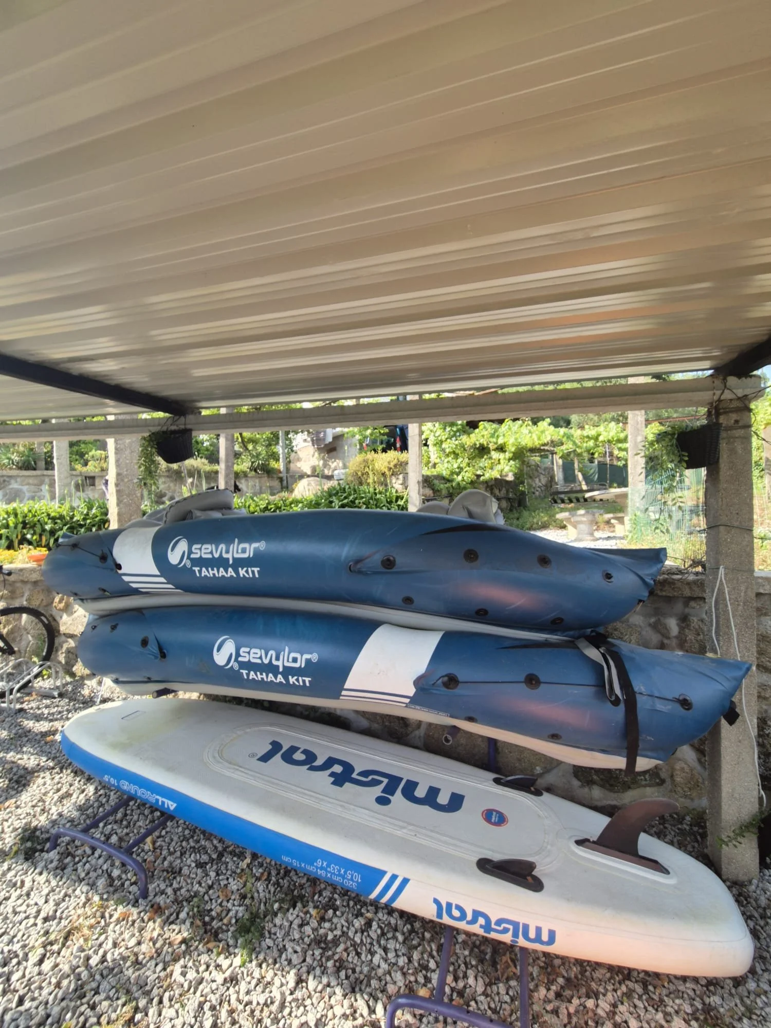 Três pranchas de stand-up paddle apoiadas contra uma parede, duas azuis e uma branca, sob uma cobertura de telhas.