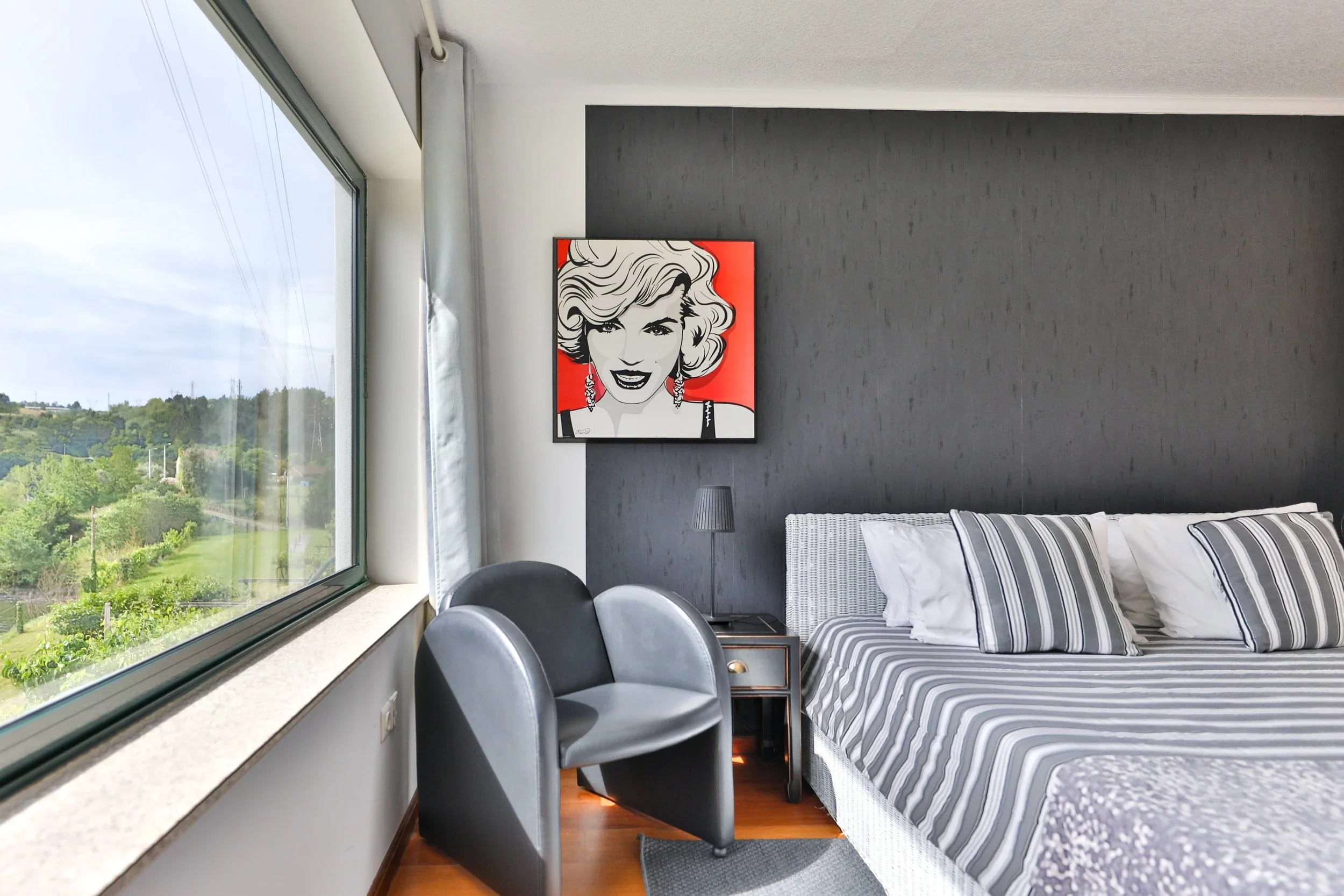 Quarto com cama de roupas listradas em preto e branco, parede escura ao fundo, quadro de retrato de mulher na parede, cadeira preta e cinza próxima à janela grande com vista para uma paisagem verde.