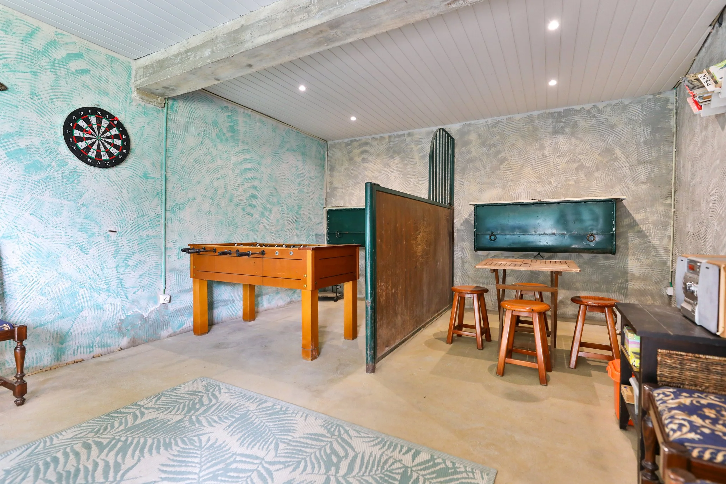 Sala de jogos com mesa de pebolim, baralho de bilhar, mesa com banquetas, arco de futebol entre as paredes, parede decorada com textura cinza e as paredes do fundo com acabamento em concreto e paredes de madeira e metal, teto de madeira com iluminaçã