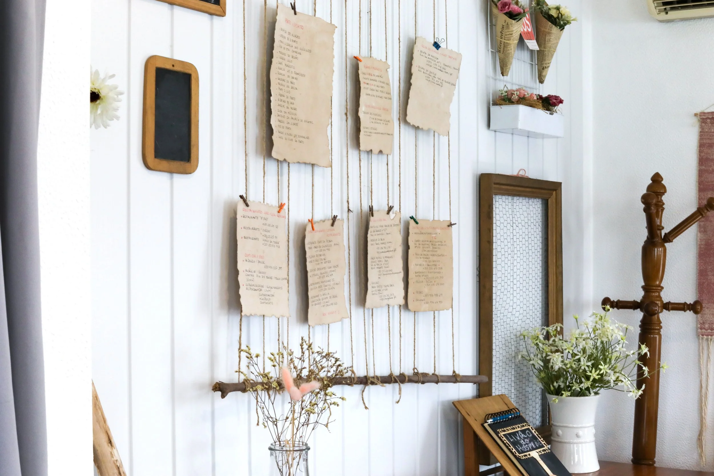 Parede decorada com folhas de papel amarradas com prendedores de roupa, algumas com textos escritos, uma moldura de madeira vazia, vasos com flores, um quadro de pensamento escrito com giz, e objetos de decoração em um ambiente aconchegante.