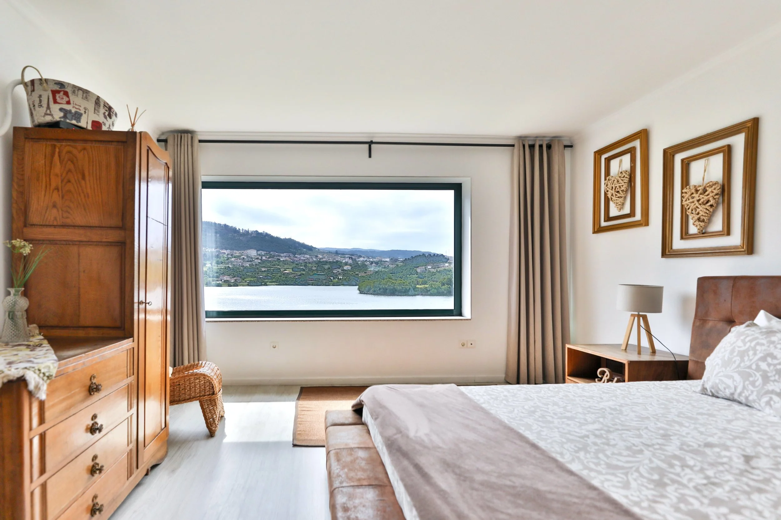 Quarto com cama, móveis de madeira, janela com vista para um lago e áreas verdes, decoração com quadros de corações de arrame, cortinas bege, luminária de mesa, e uma cabeceira de cama marrom.