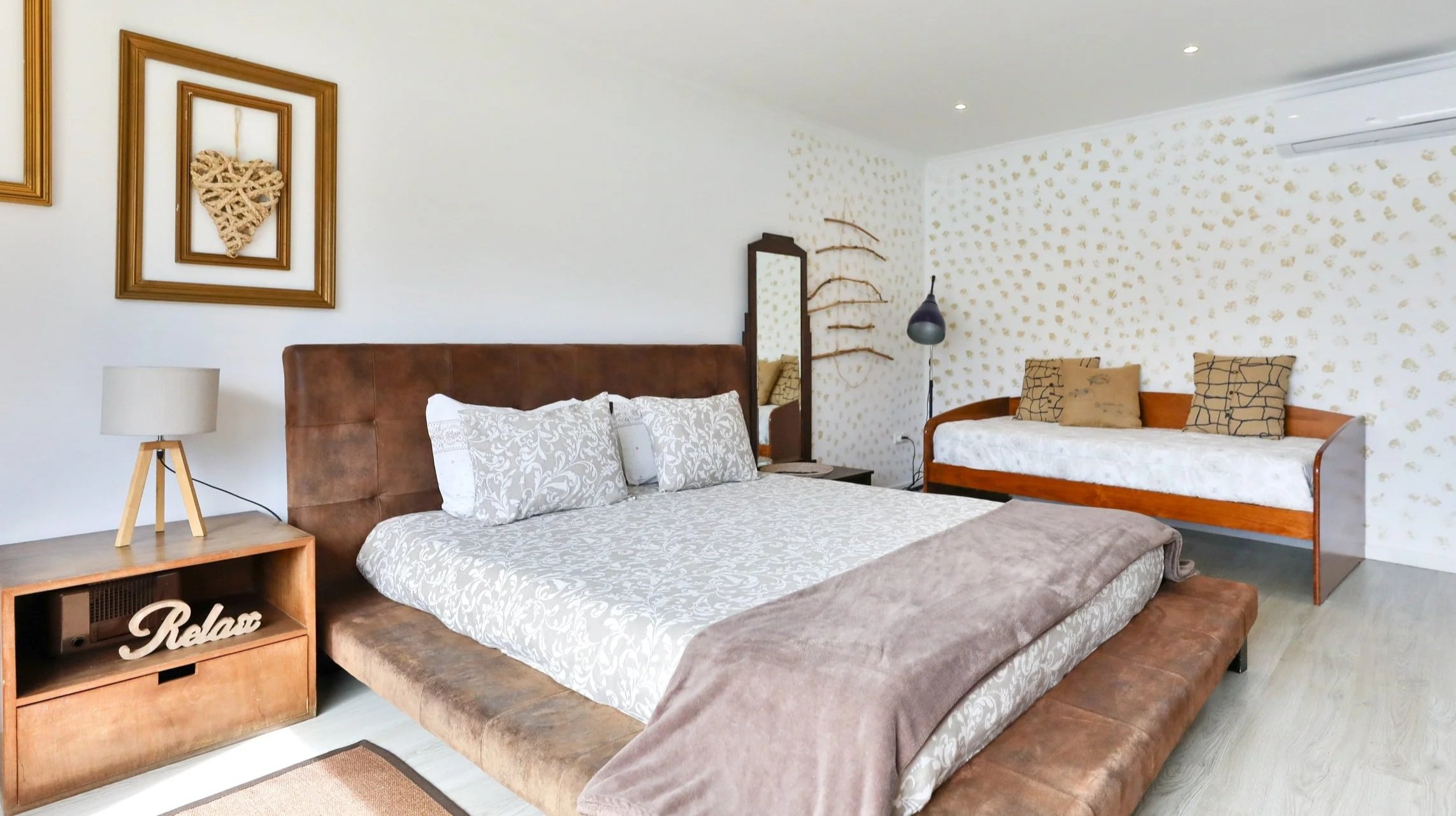 Quarto com cama de casal com cabeceira de estofado marrom, travesseiros brancos e roupa de cama com estampa floral, criado-mudo de madeira com luminária de mesa e decoração de quadros na parede, poltrona de madeira com almofadas decorativas, espelho 