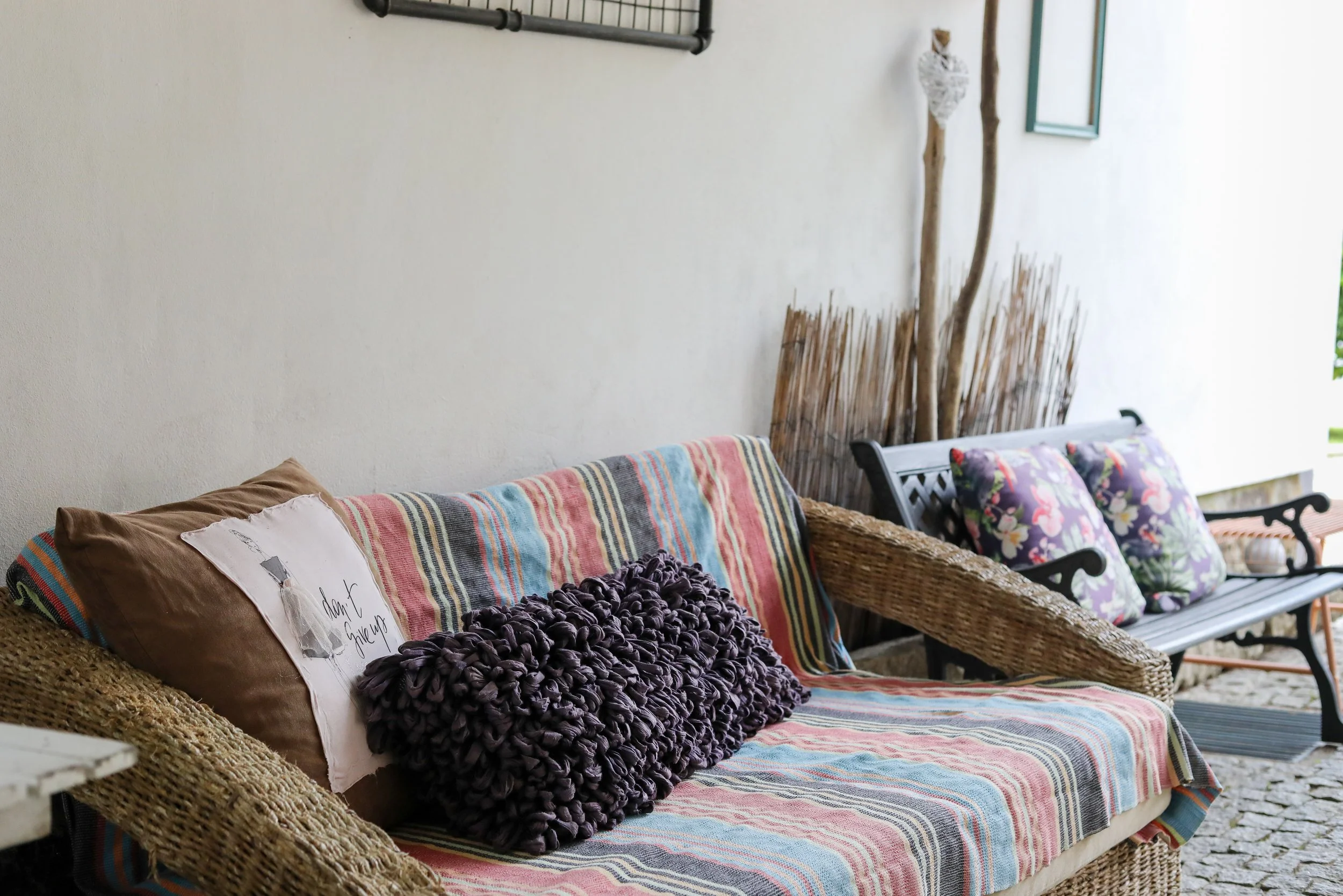 Sala de estar com sofá de vime, almofadas decorativas, uma poltrona de vime com almofadas de flores, e decoração rústica com árvores secas e cercado de galhos ao fundo.