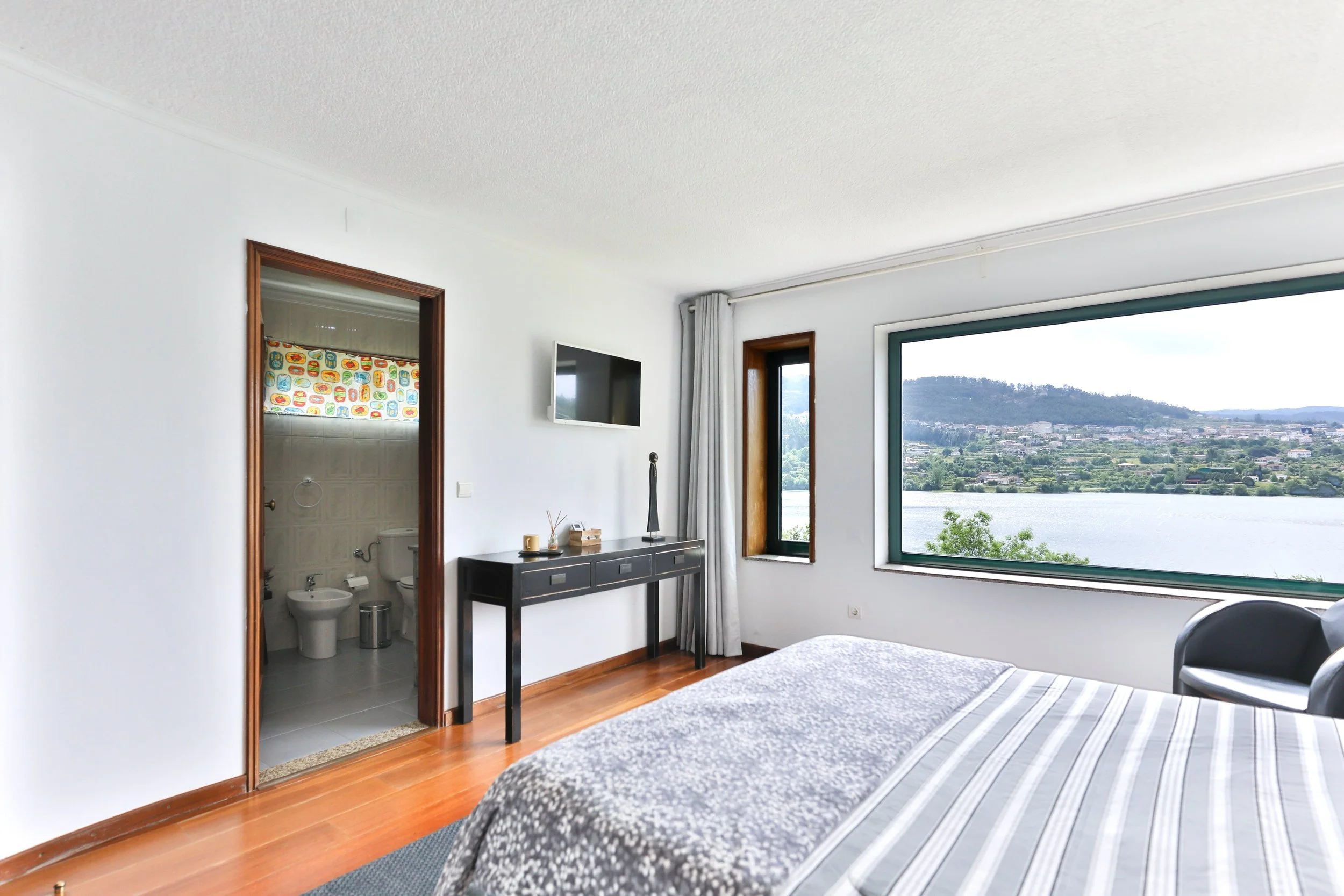 Quarto de hotel com vista para um lago, contendo cama de casal, televisão na parede, janela grande e uma porta que leva ao banheiro.