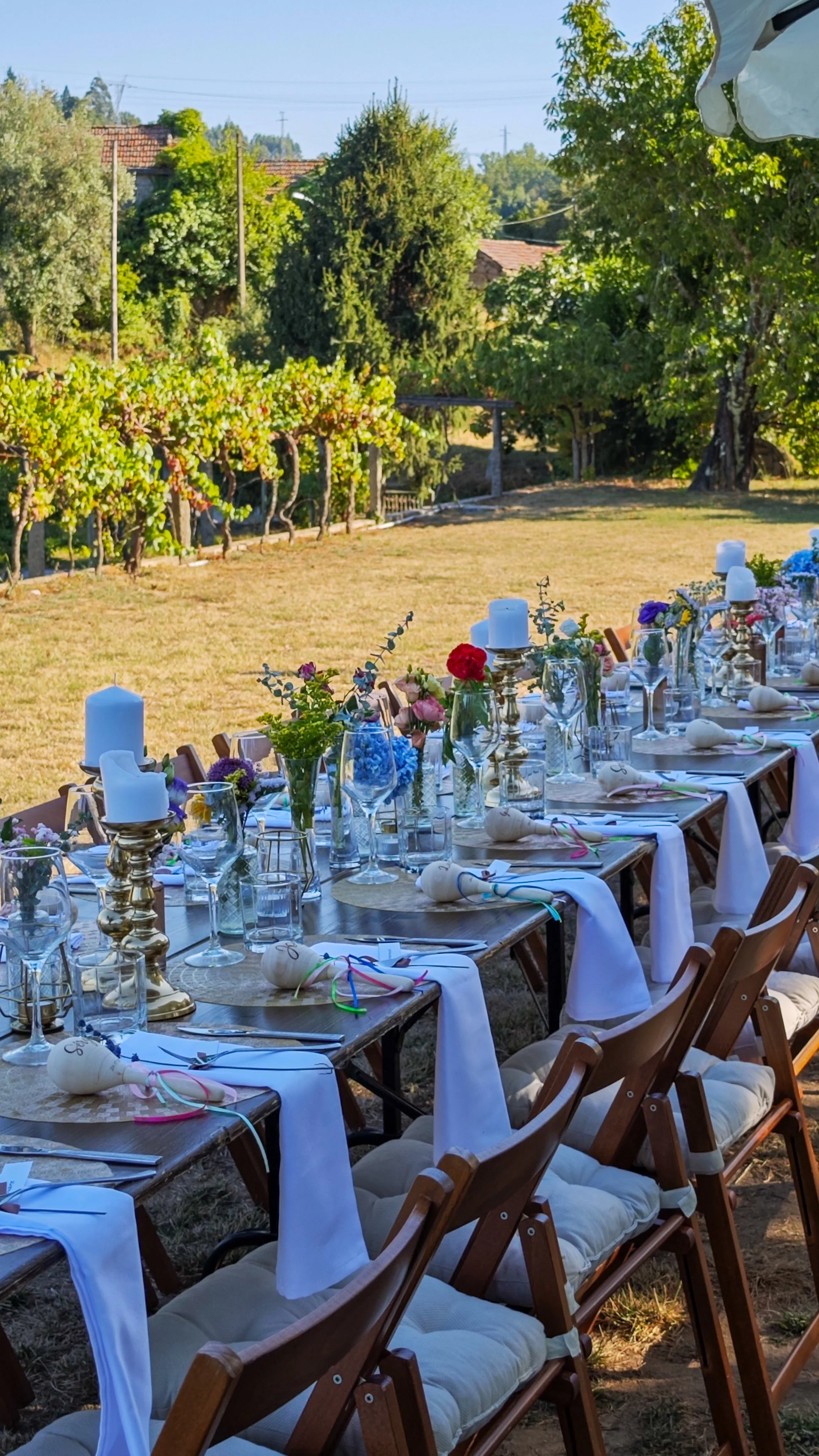 Mesa de jantar decorada com velas, flores e guardanapos em um ambiente ao ar livre, com árvores e vegetação ao fundo.