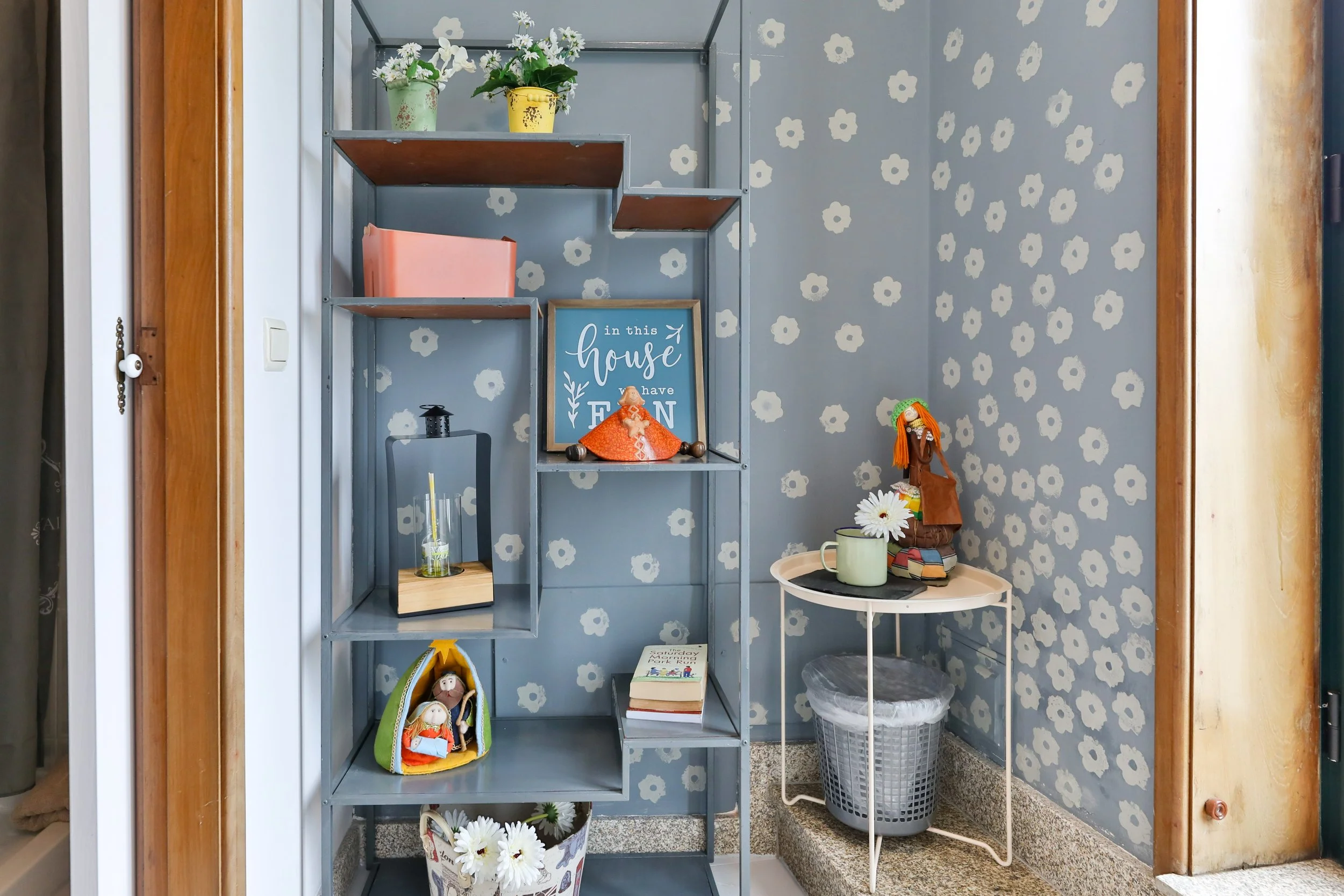 Requintado espaço de interior com paredes azuis com estampa de flores brancas, uma estante de metal com prateleiras de madeira exibindo objetos decorativos, incluindo bonecos de palha, livros, uma moldura com frase e vasos de flores, além de uma mesa