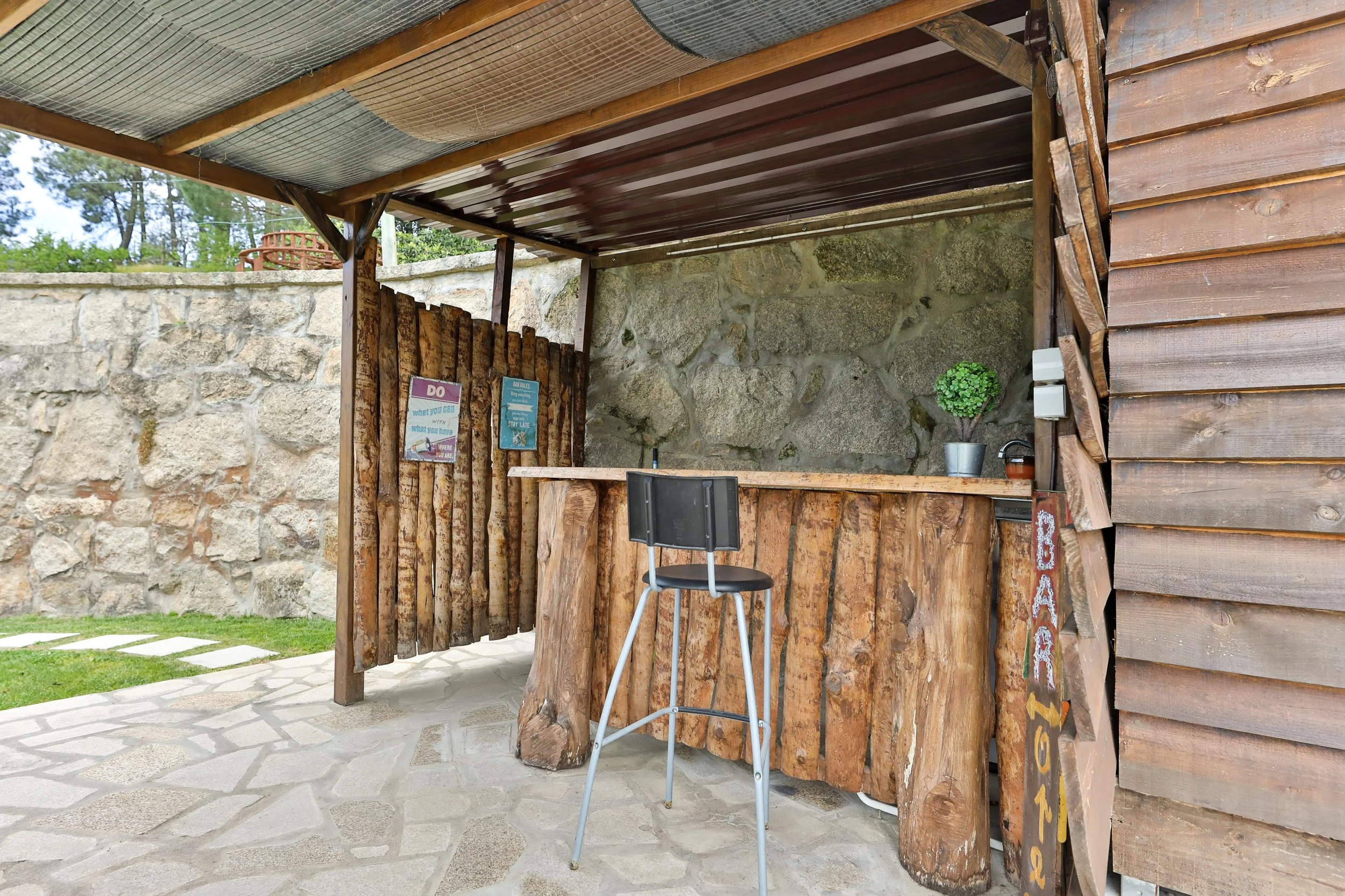 Bar de madeira ao ar livre com tampo de madeira, cadeira preta e parede de pedra ao fundo, decorado com um vaso de planta, sinais coloridos e uma placa com a palavra 'Bar' na lateral.