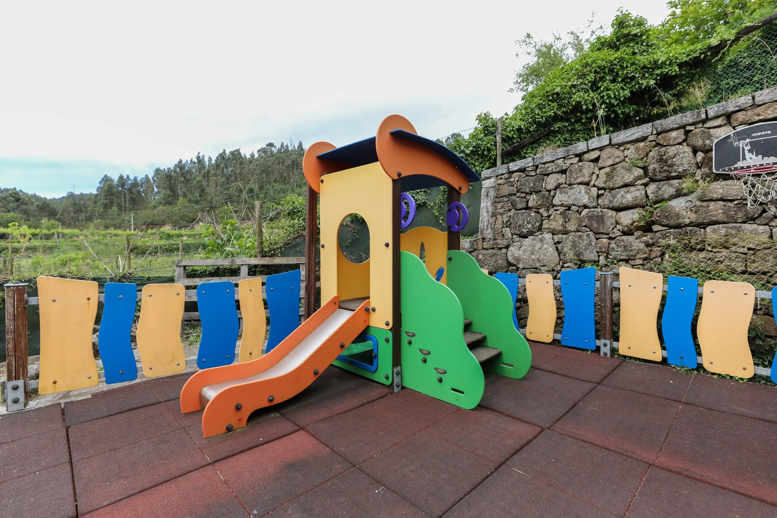 Playground com escorregador colorido, cercado por painel de madeira e pano de fundo com muro de pedra e natureza ao redor.