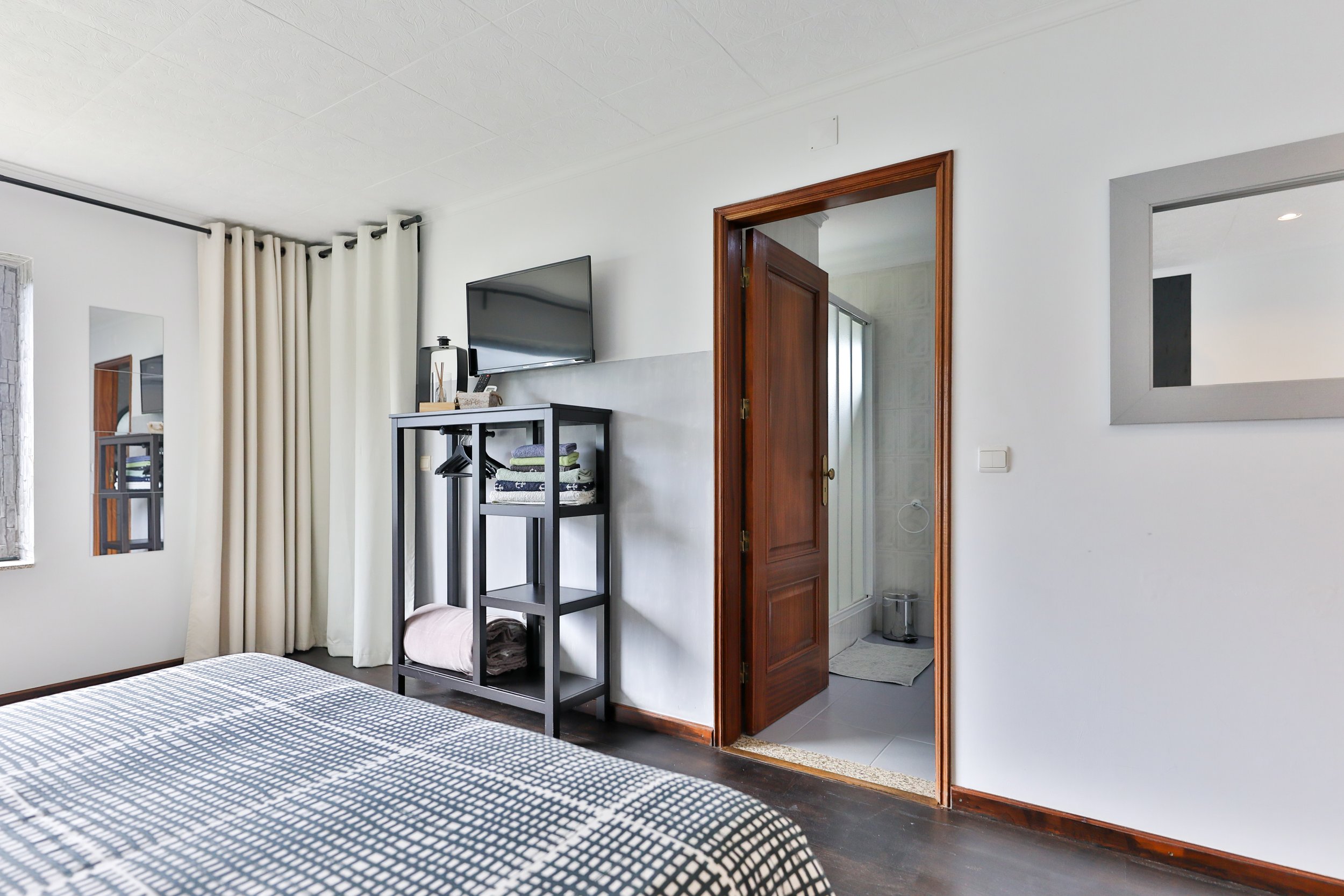 Quarto com cama de lençol listrado, janela com cortinas brancas, espelho na parede, televisão e uma estrutura preta com roupas e toalhas, porta de entrada para o banheiro ao lado.