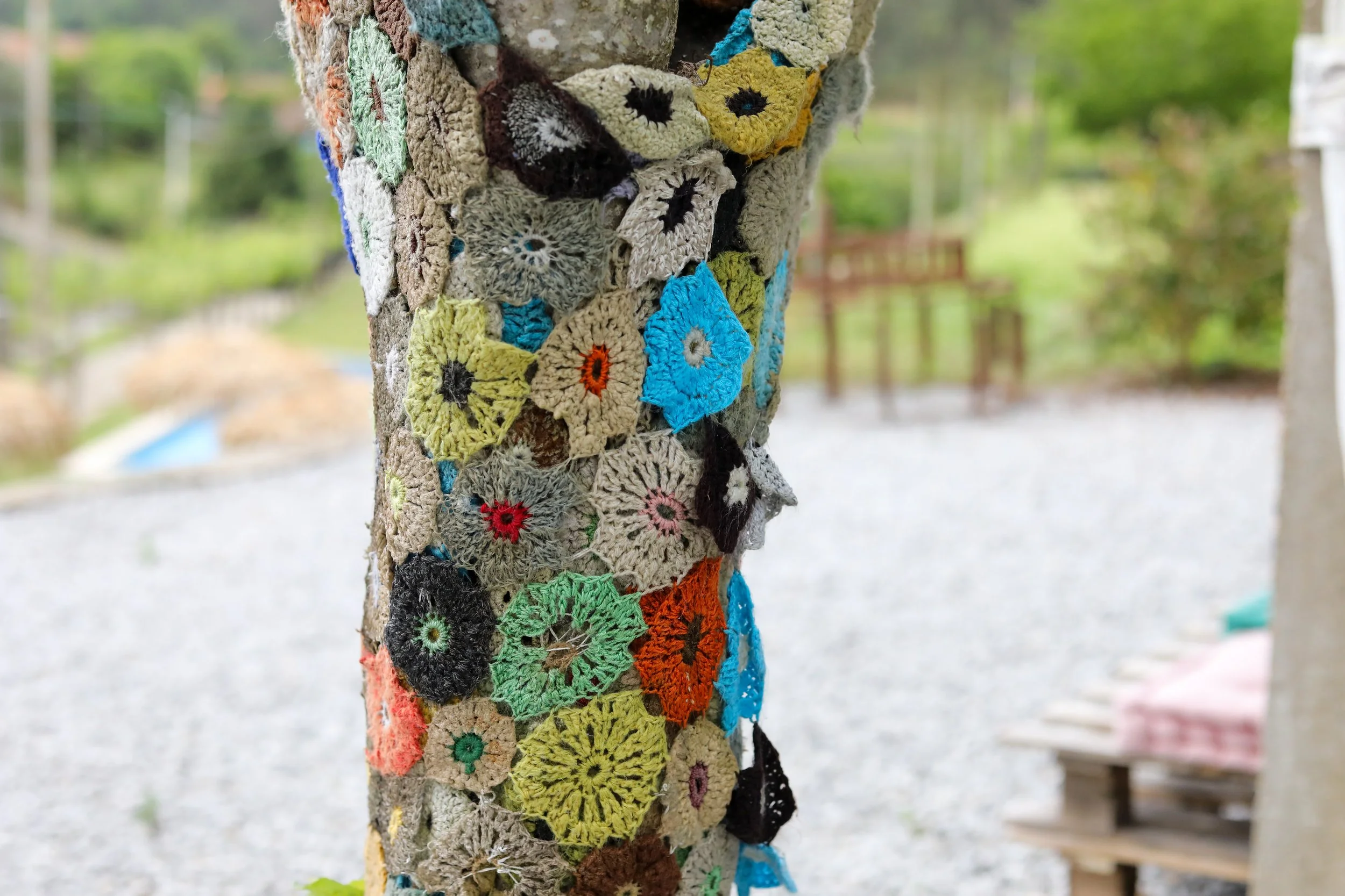 Árvore decorada com crochê colorido, secções variadas e detalhes em diferentes cores, numa área ao ar livre com bancos e vegetação ao fundo.