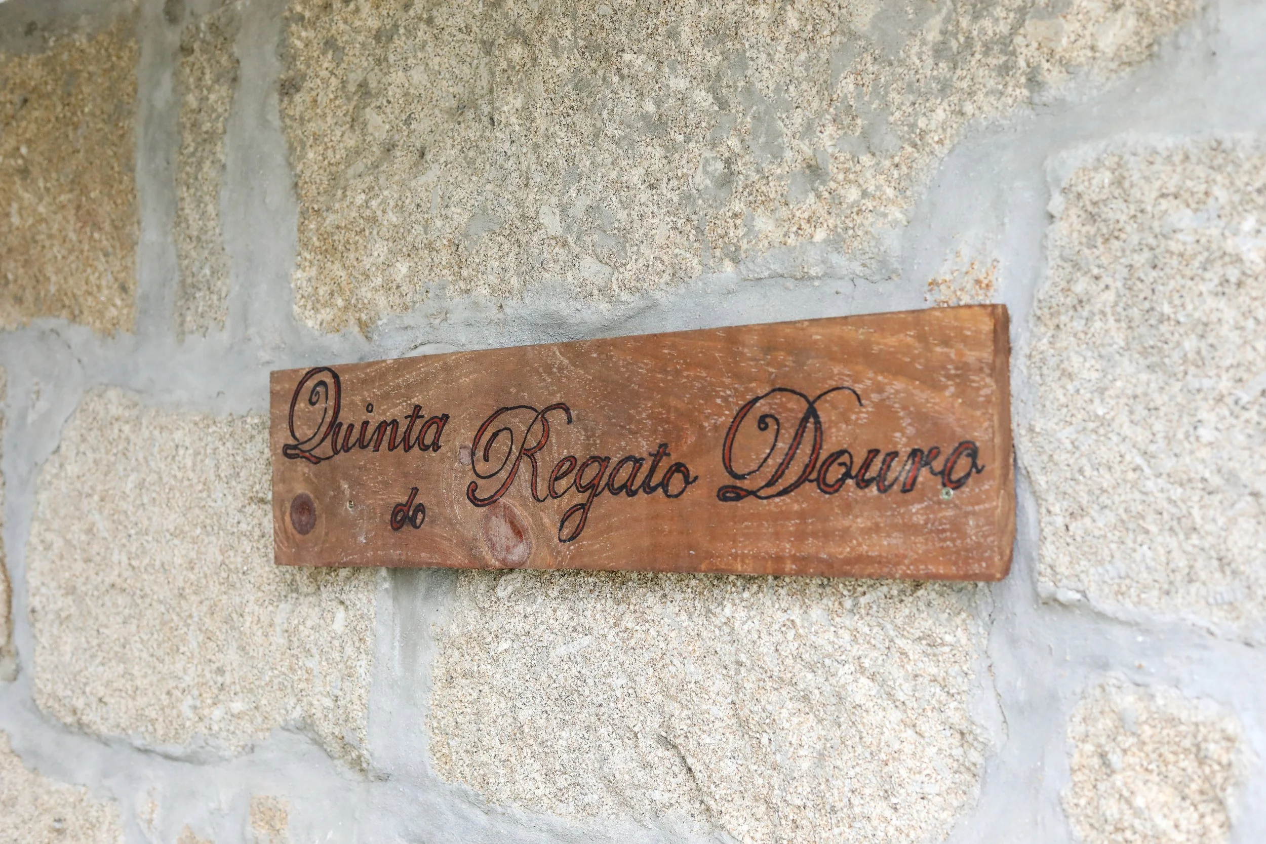 Placa de madeira com escrita em português 'Quinta Regato Douro' instalada em uma parede de pedras.