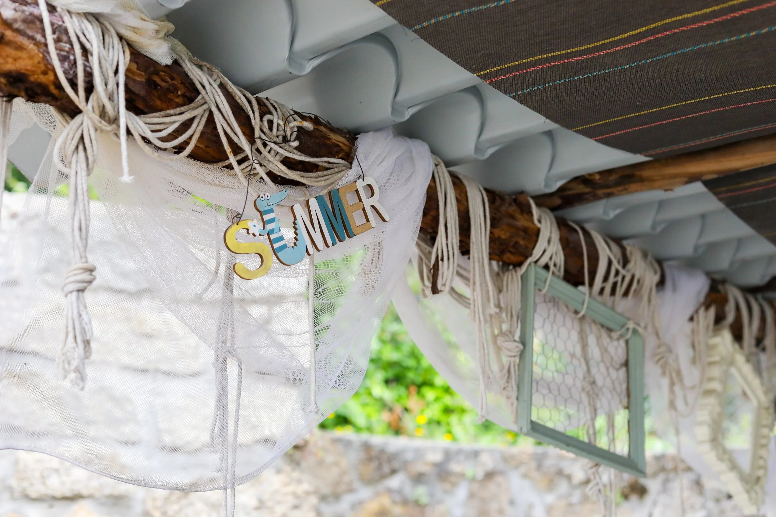 Decoração de verão com uma inscrição pendurada que diz 'SUMMER' ao lado de um desenho de sereia, tudo em ambiente ao ar livre com fundos de árvores e céu verde.