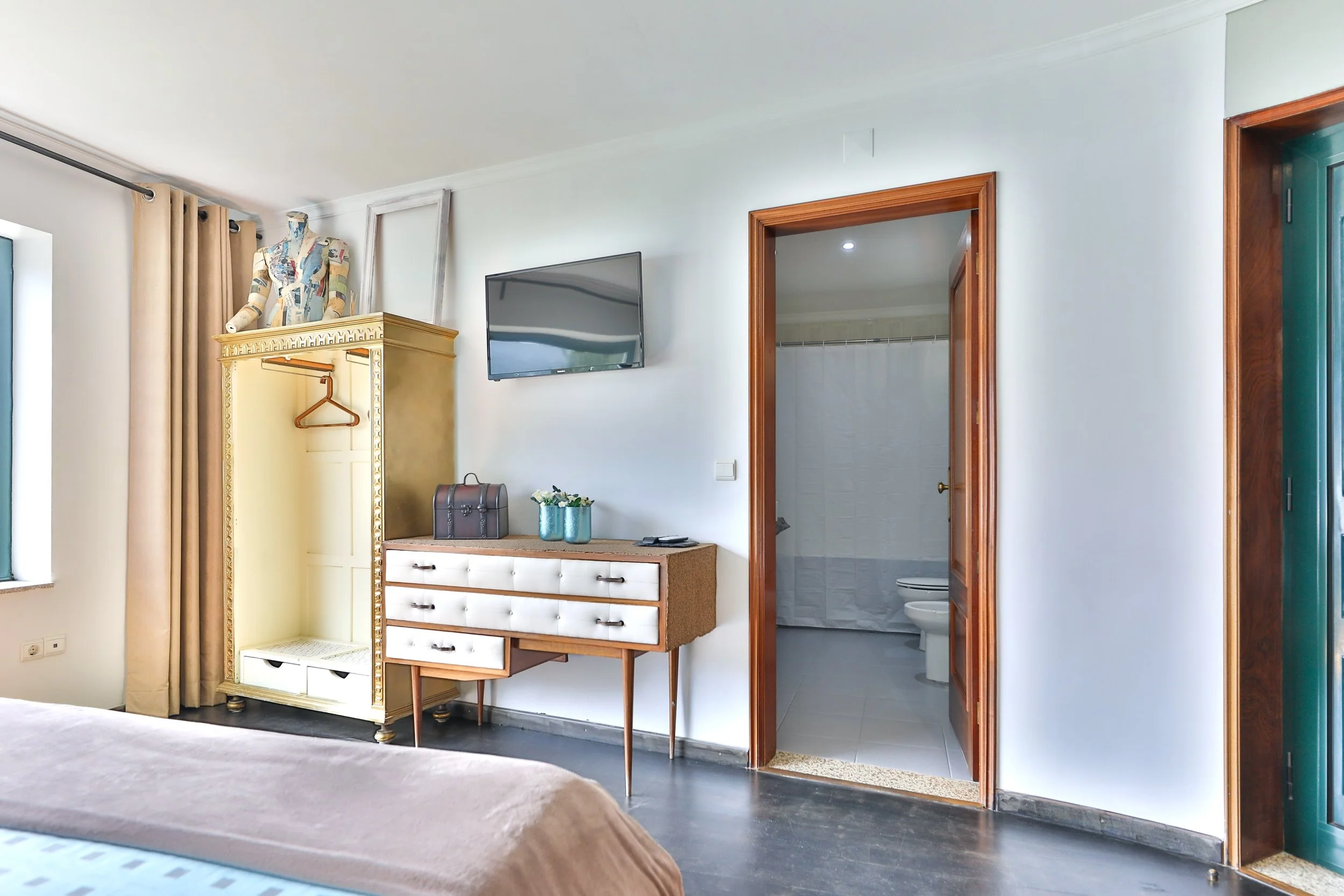 Quarto com armário amadeirado, televisão de parede, uma cômoda vintage, banheiro ao fundo com pia e vaso sanitário, janela com cortina bege, cama parcialmente visível com colcha cinza e marrom.