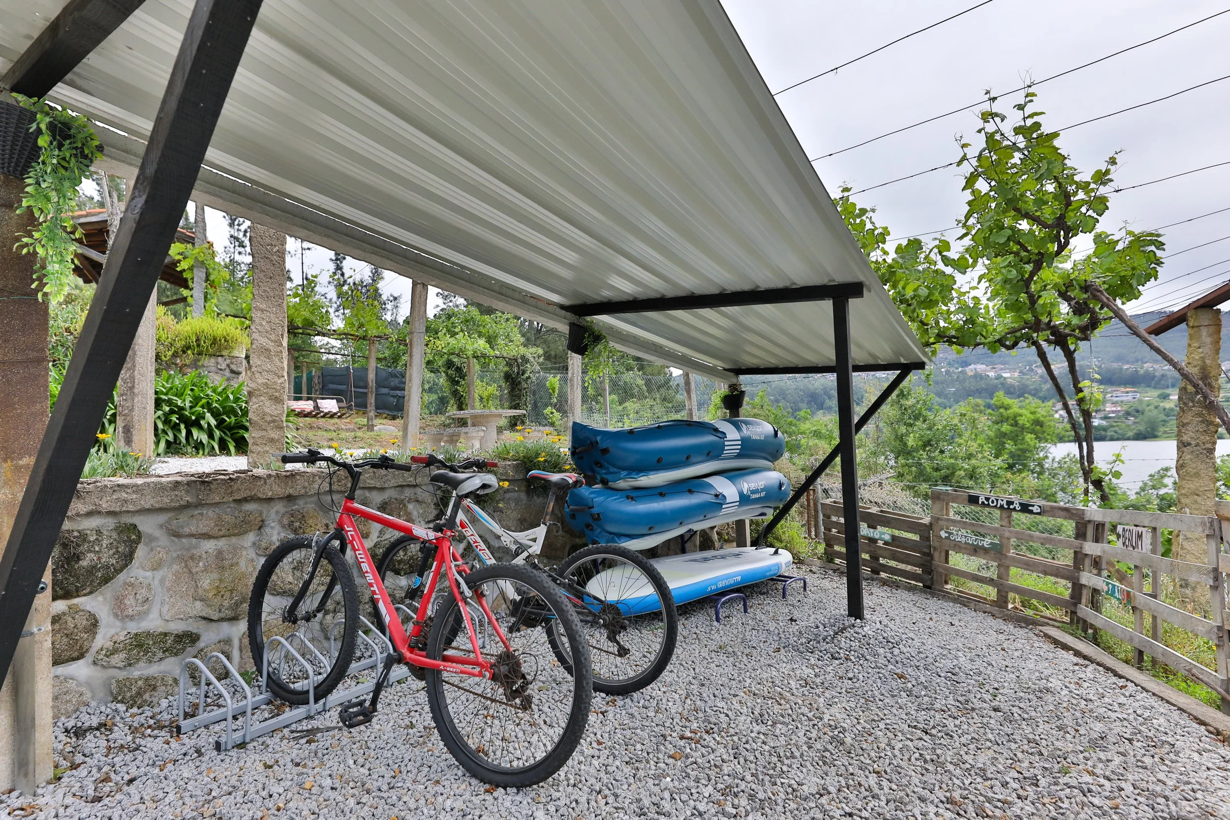 Estacionamento ao ar livre com duas bicicletas, uma vermelha e uma branca, apoiadas em um suporte de metal, e uma cobertura de metal sobre elas. Ao fundo, há um muro de pedra, plantas, árvores, uma cerca de madeira e uma paisagem com rio e vegetação.