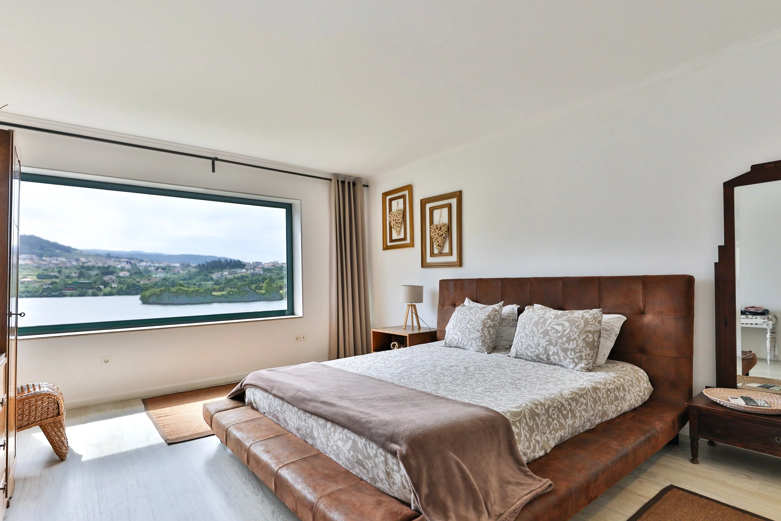Quarto de hotel com cama de casal, travesseiros decorativos em fundo branco, janela grande com vista de rio, decoração com quadros de coração na parede, cortinas bege, tapete e móveis de madeira.