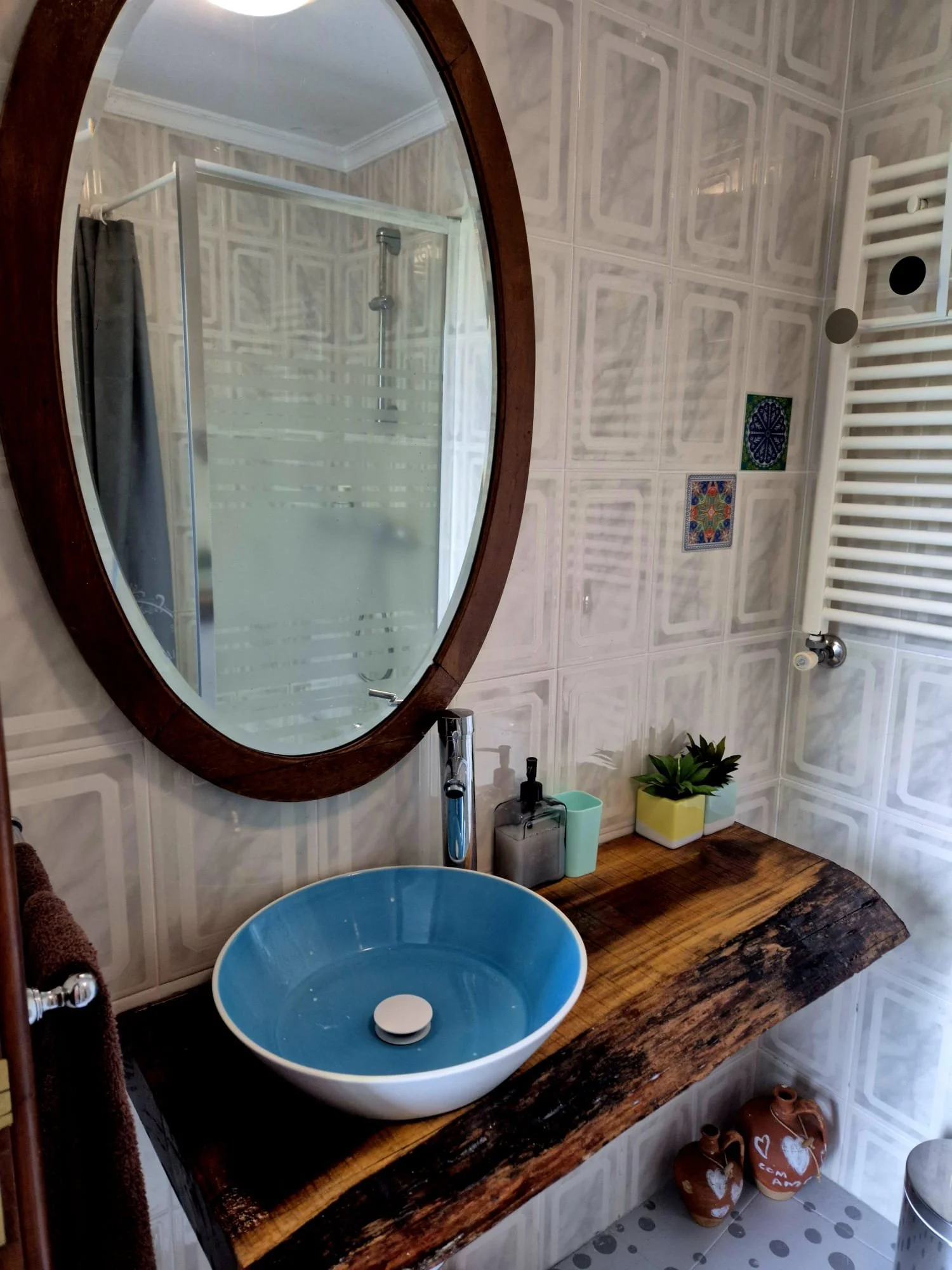 Banheiro com parede de azulejos, espelho oval com moldura de madeira, pia de sobrepor azul, bancada de madeira rústica, vaso de plantas, sabonete, escova de dente e outros itens de higiene, toalha marrom e vaso decorativo com corações.