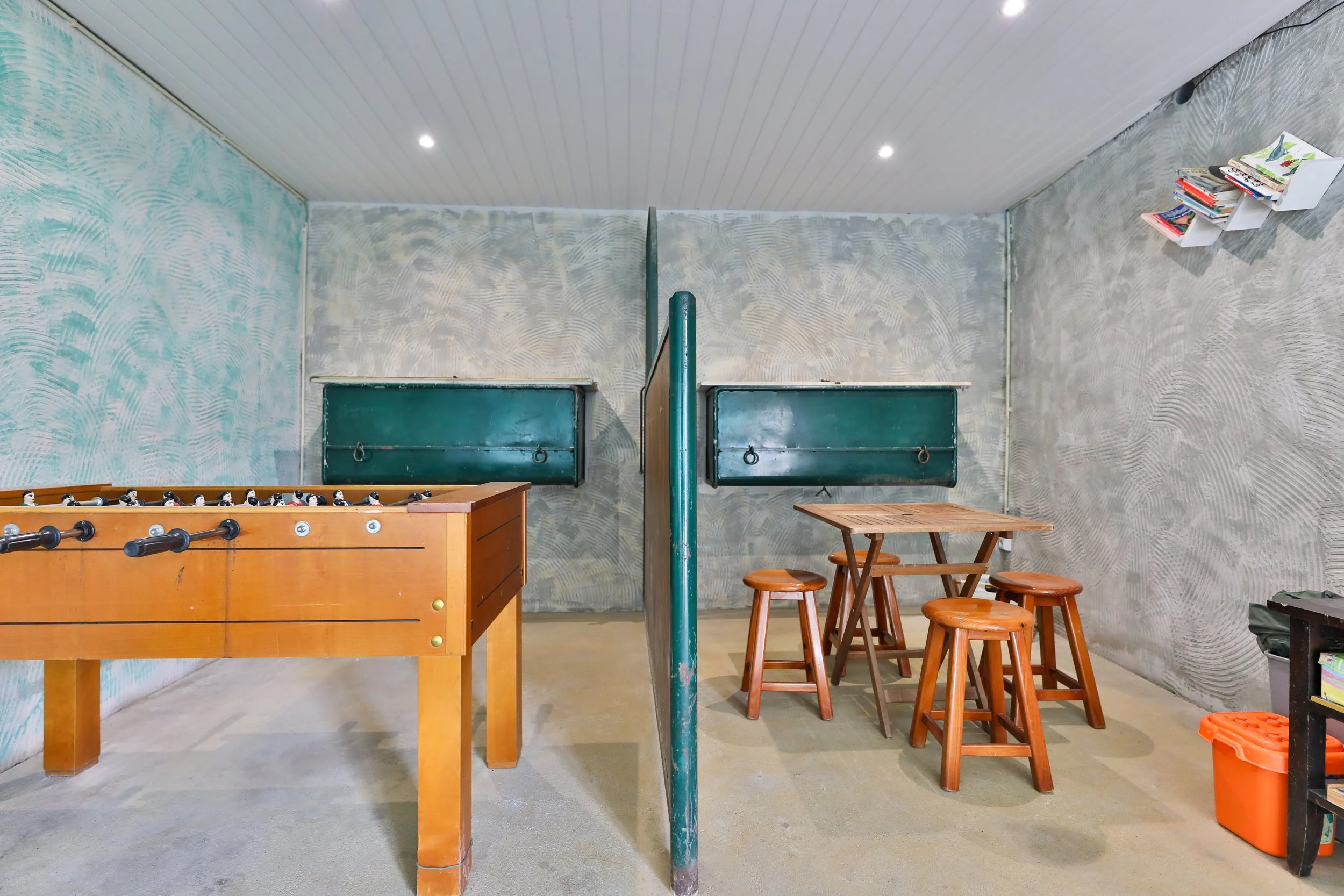 Sala de jogos com mesa de pebolim, mesa de madeira com bancos, paredes com textura, prateleiras de livros na parede, piso de cimento e teto branco com iluminação