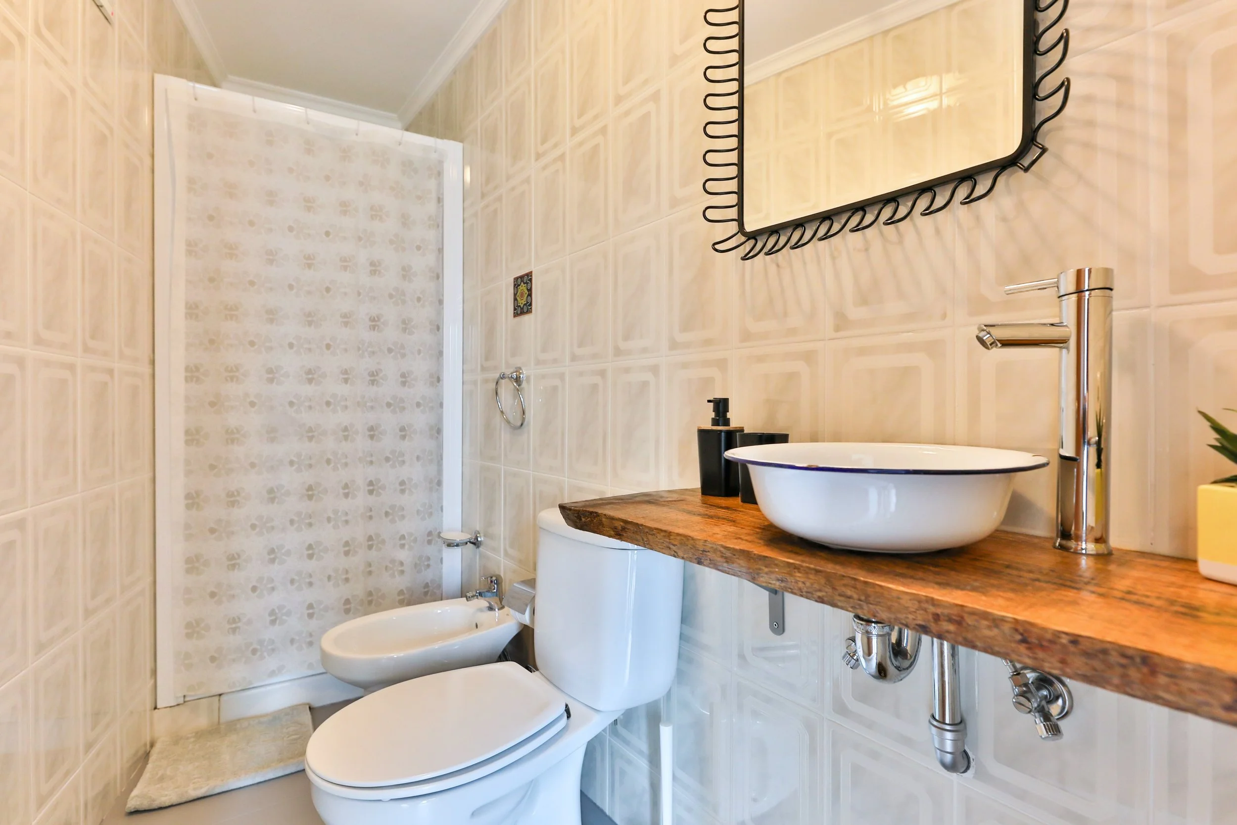 Banheiro com vaso sanitário, bidê, pia de apoio de madeira com cuba de sobrepor, espelho com moldura decorativa, armário com sabonete líquido e planta em vaso na bancada.