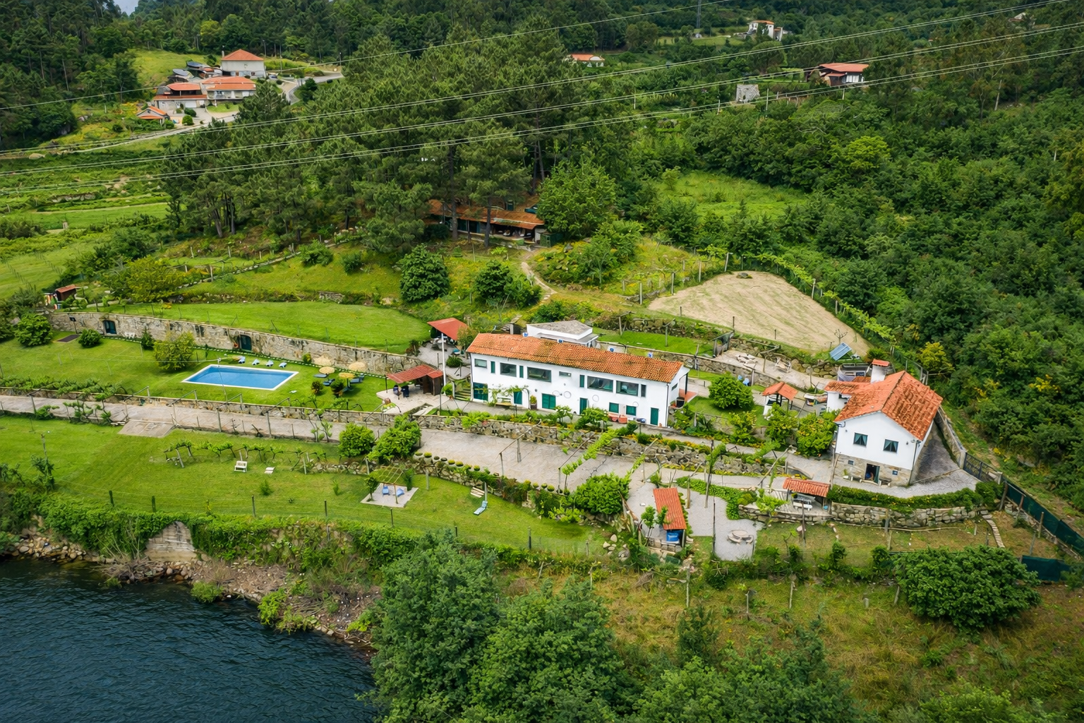 Vista aérea de uma propriedade com casas brancas, piscina, jardim bem cuidado, muro de pedra, árvores e caminhos, localizada próxima a um corpo de água e cercada por área verde e floresta.