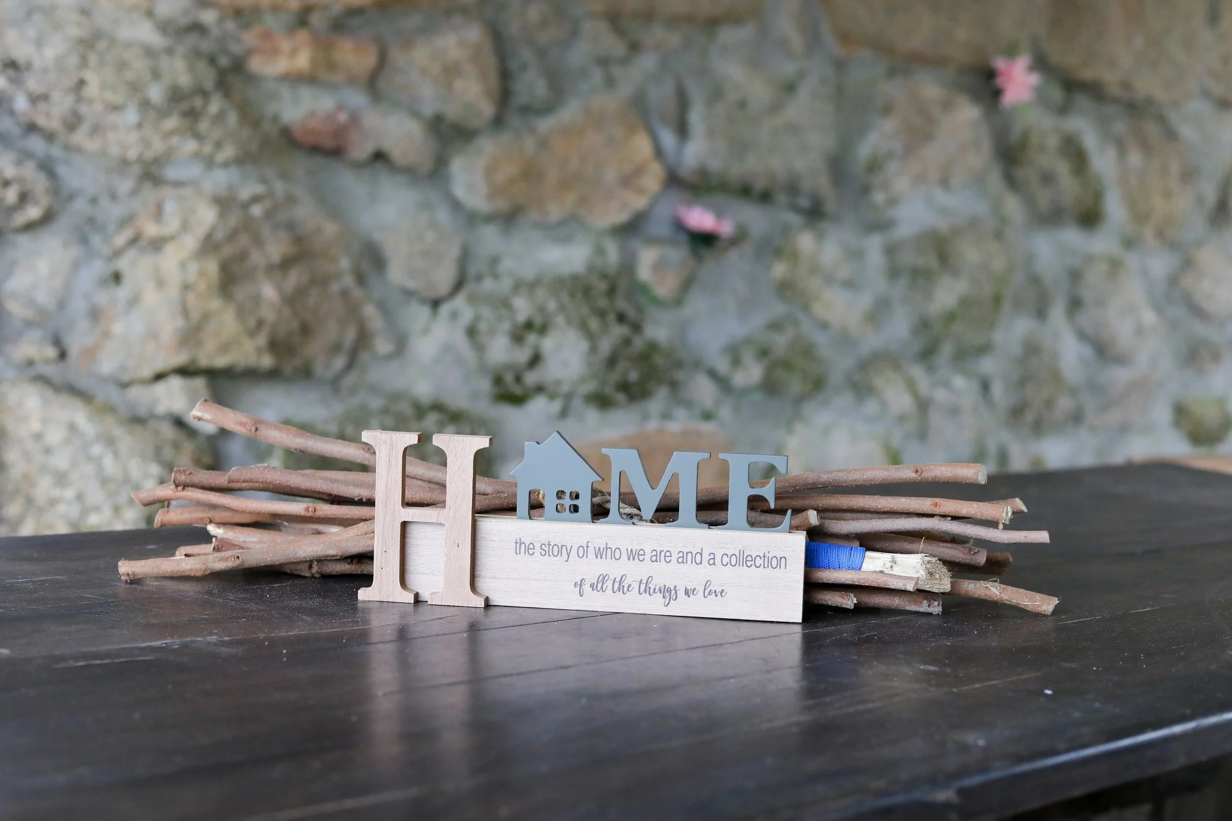 Decoração de mesa com objetos de madeira formando a palavra 'HOME', com uma frase que diz 'a história de quem somos e uma coleção de todas as coisas que amamos', ao fundo uma parede de pedra.
