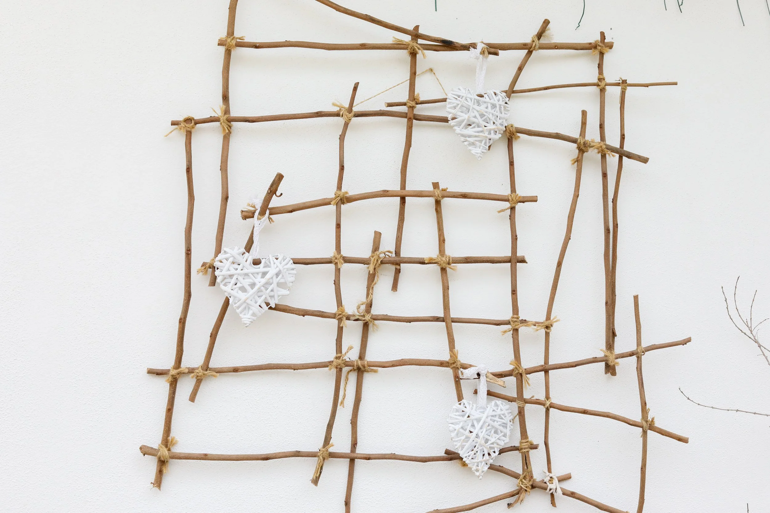 Estrutura de galho de madeira presa com lascas de madeira, decorada com quatro corações brancos de enfeite pendurados com tiras de tecido branco, instalada na parede branca.