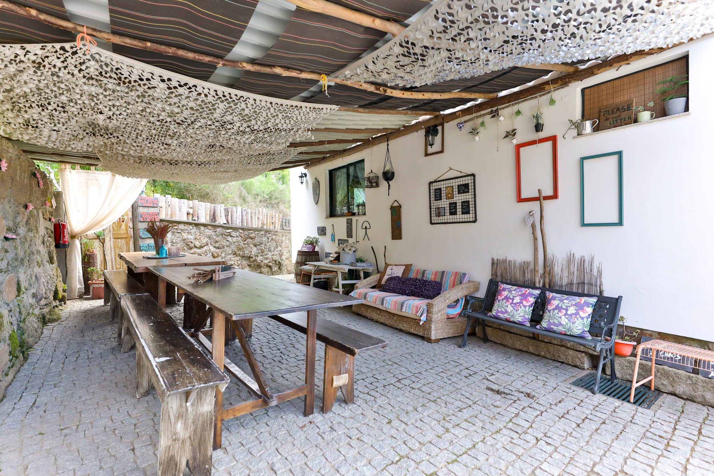 Escritório ao ar livre com mesa de madeira, bancos de tronco, sofá com almofadas, bancos de ferro com almofadas florais, paredes decoradas com quadros, plantas e objetos decorativos, teto com tecidos e madeiras, piso de pedra.