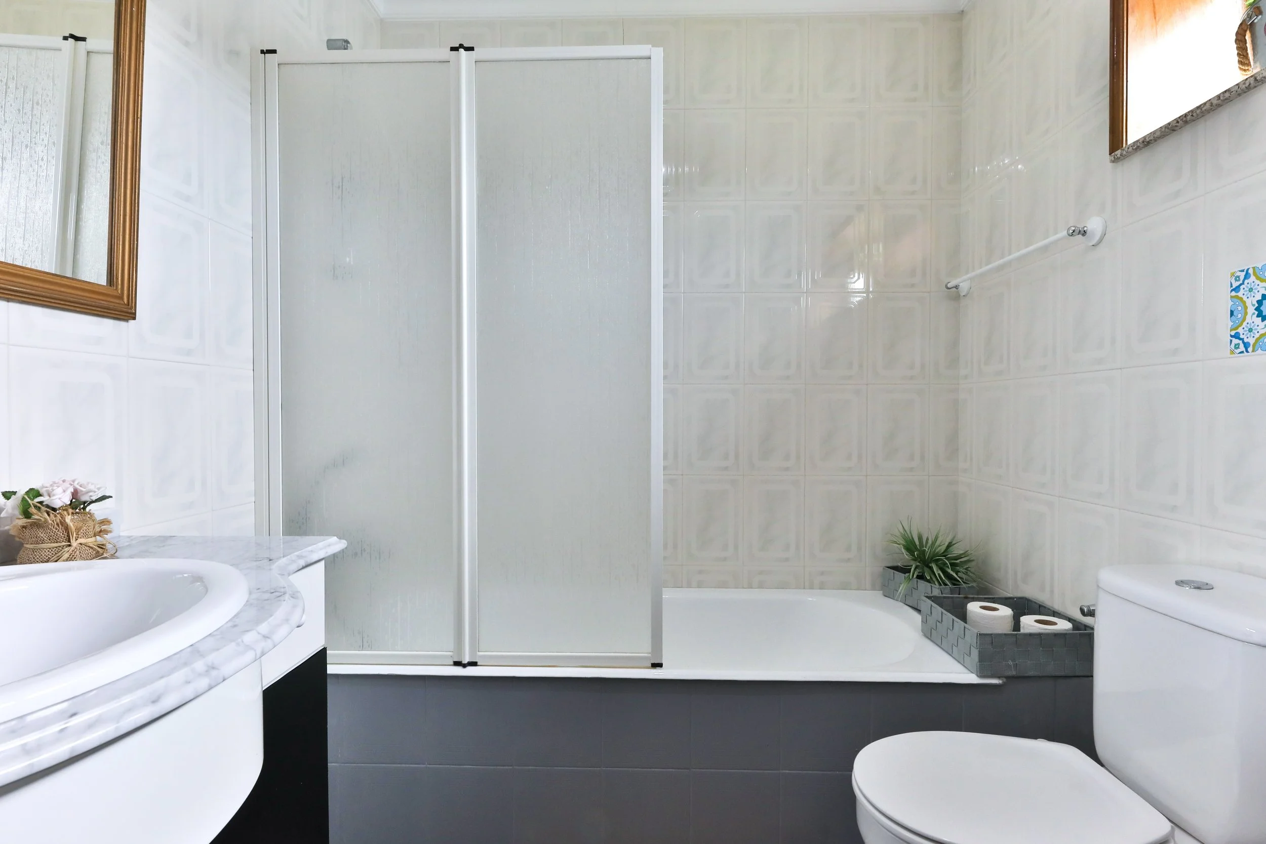 Banheiro com pia, espelho com moldura dourada, vaso sanitário, banheira, divisória de vidro fosco, plantas decorativas, toalha na parede, caixas com papel higiênico, paredes revestidas com azulejos brancos e piso cinza.