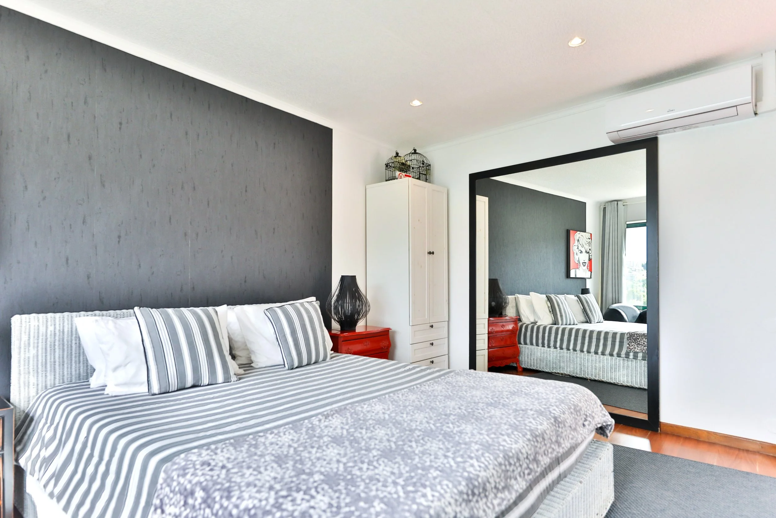 Quarto com cama de roupa de cama listrada em azul e branco, duas mesas de cabeceira vermelhas, uma luminária preta, armário branco e espelho grande refletindo a cama e decoração moderna