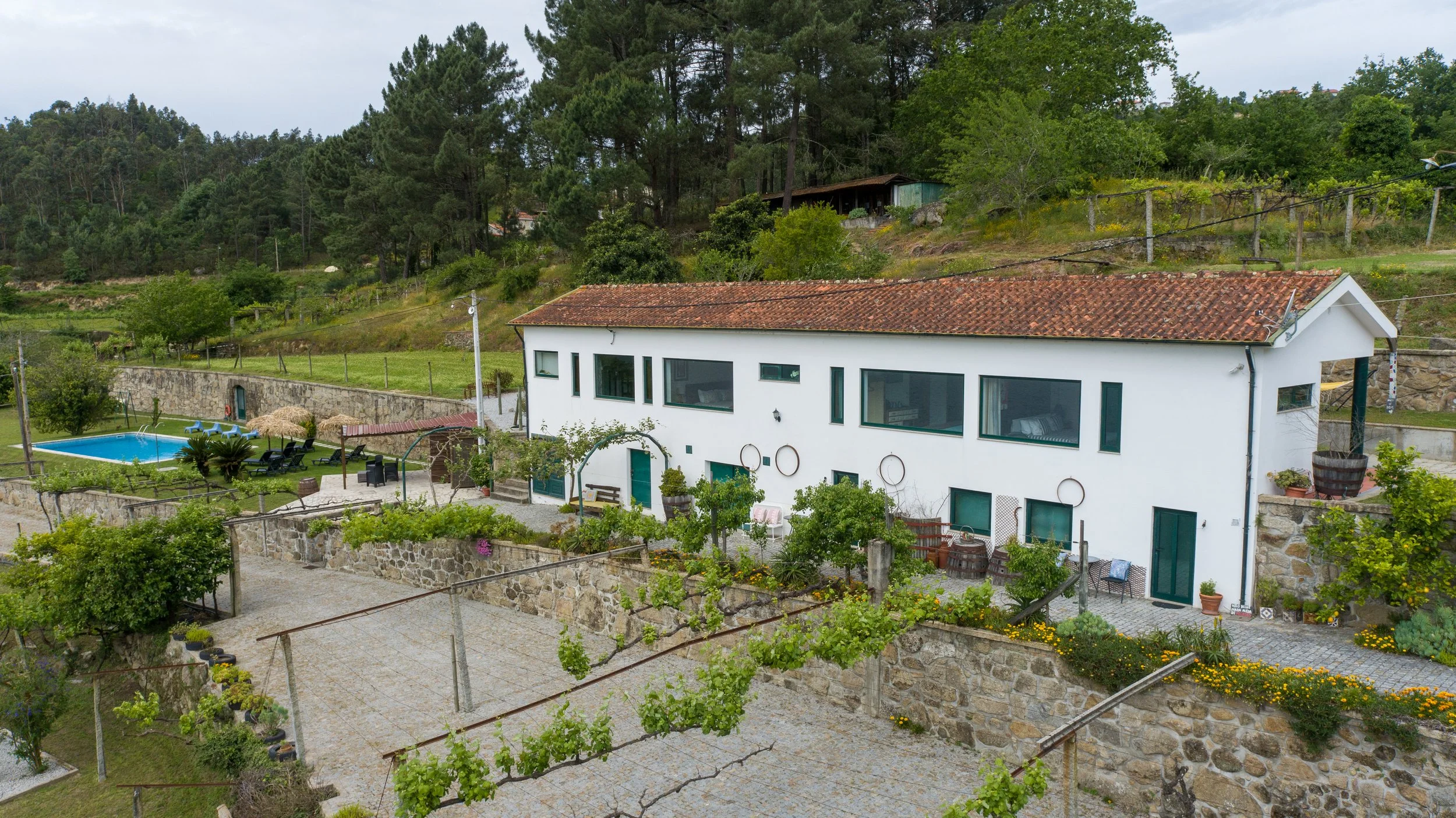 Casa branca com janelas grandes e telhado de telhas vermelhas, jardim com árvores e plantas, piscina ao ar livre, e área de lazer com mobiliário, situados em uma área rural com árvores e colinas verdes ao fundo.
