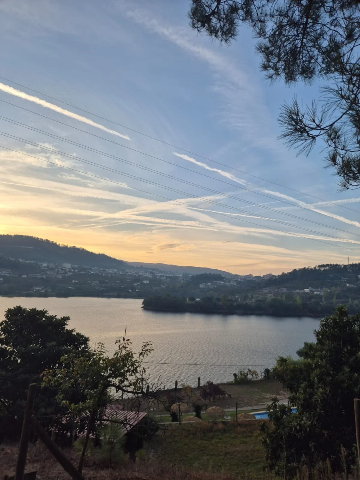 Paisagem natural ao entardecer, com rio, árvores, céu com nuvens e linhas de avião.