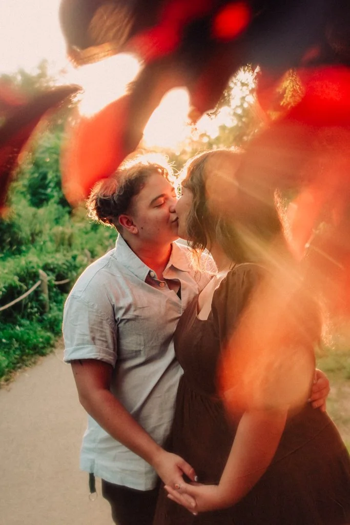 A couple is kissing outdoors during sunset, with a blurred couple in the foreground and a green natural background.
