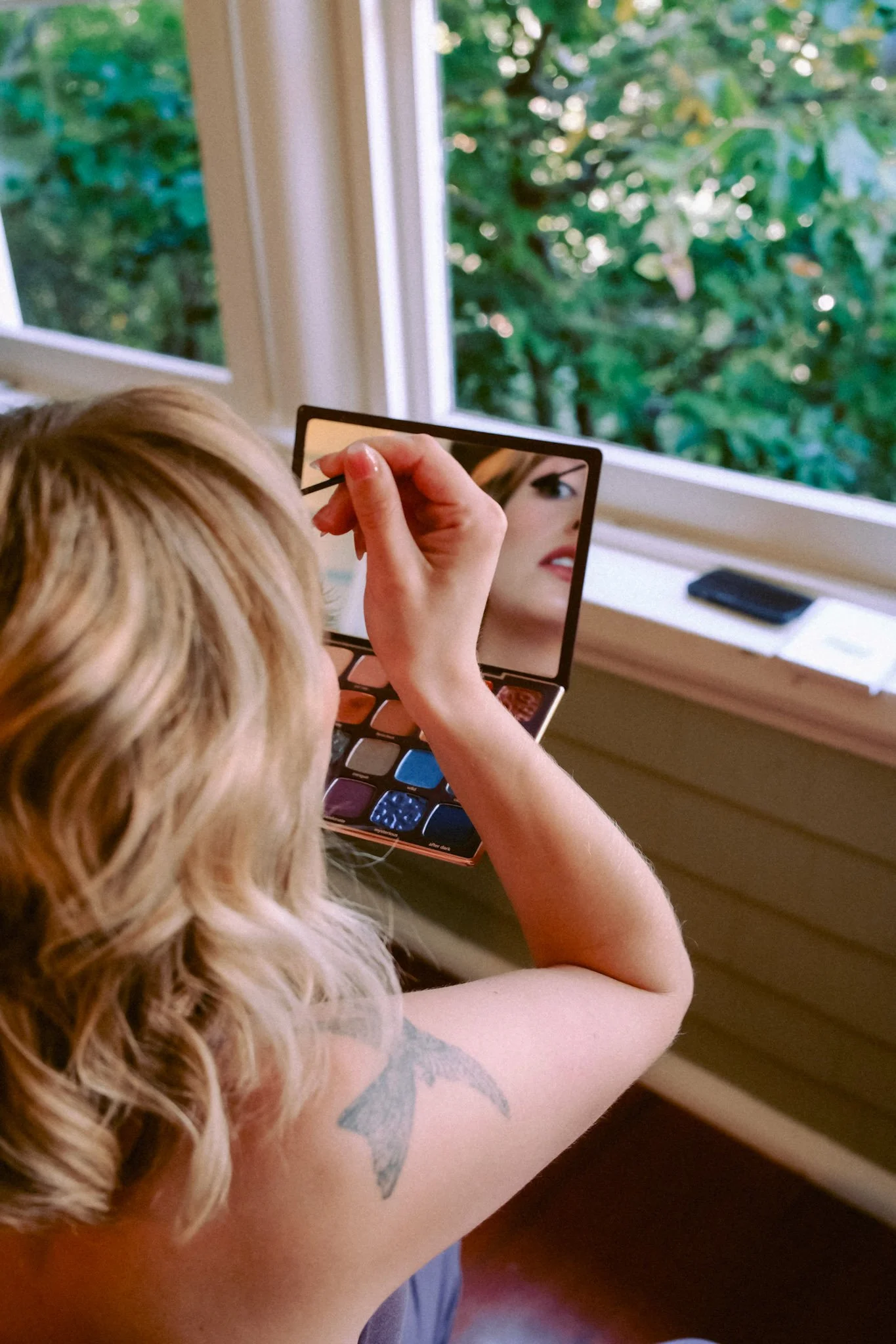 A woman applying makeup while looking into a compact mirror, near a window with a view of green foliage outside.
