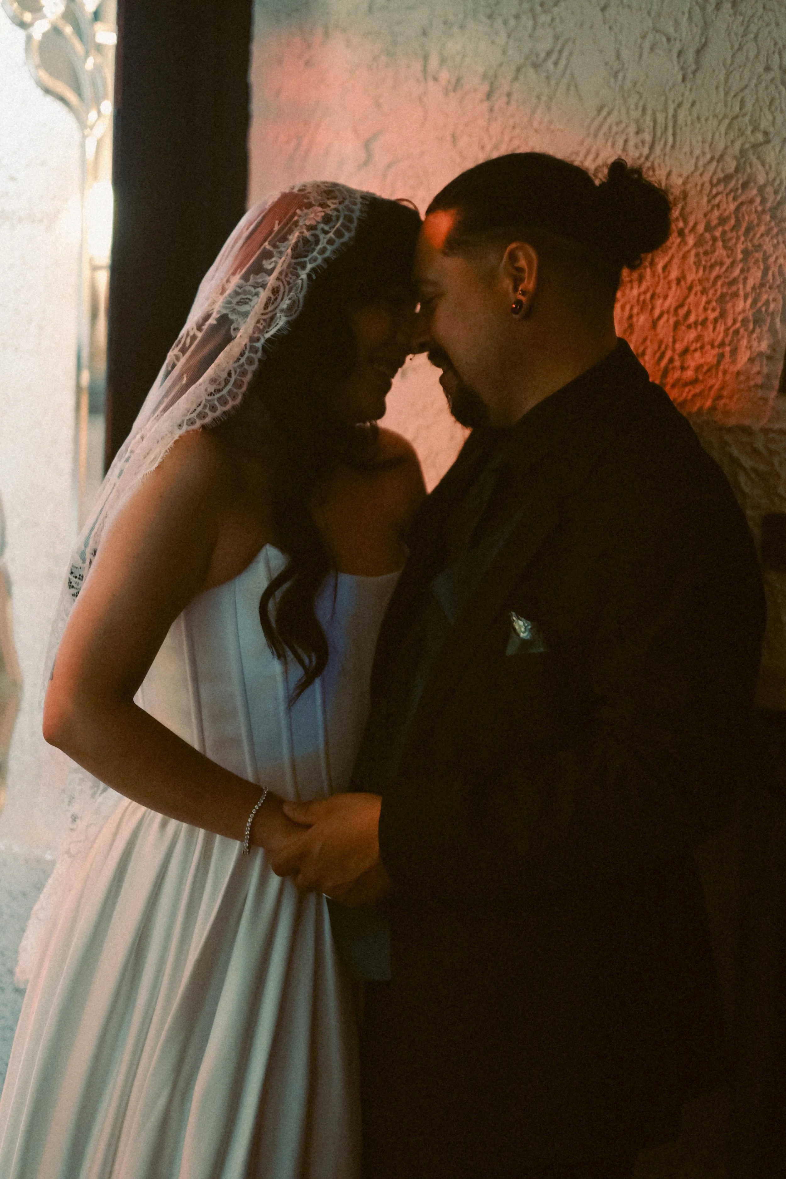 A couple is sharing a close, intimate moment with their foreheads touching, standing in front of a textured wall near a window with white curtains.