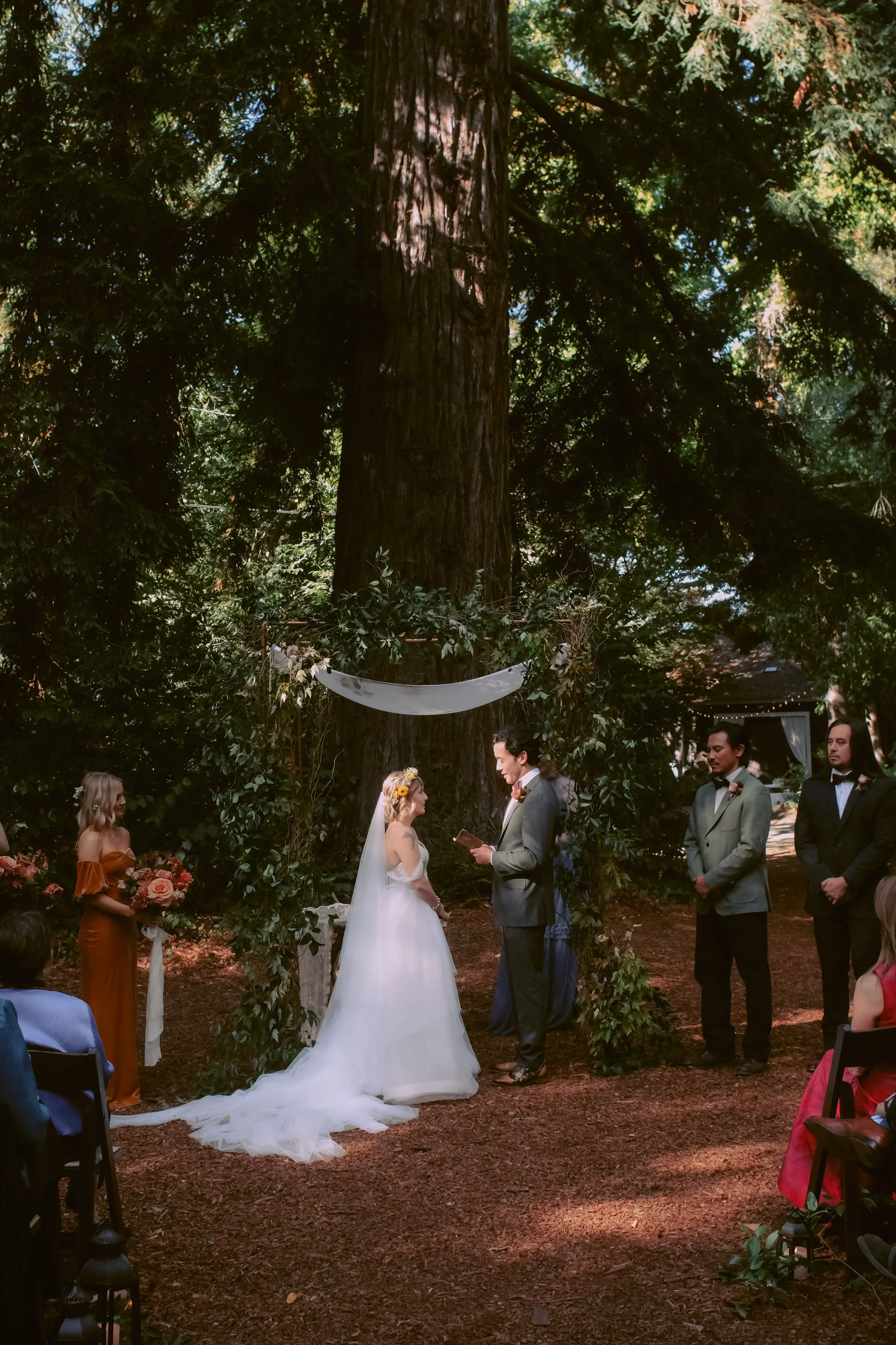 A couple getting married outdoors underneath a large tree, with a officiant reading during the ceremony. The bride in a white wedding dress with a veil and a flower crown, and the groom in a gray suit. Bridesmaids and groomsmen stand nearby, with gue