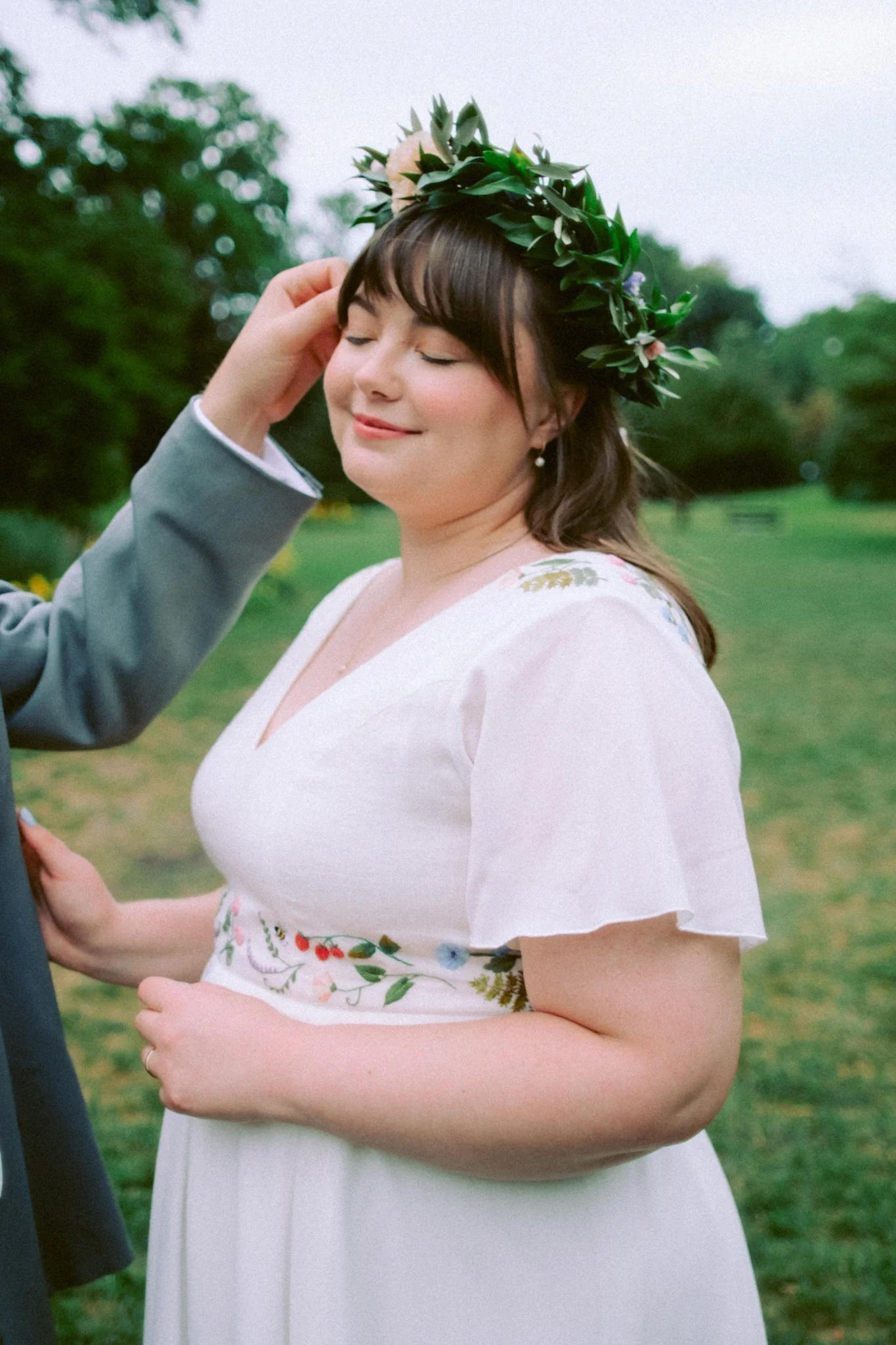 A woman with brown hair wearing a white dress with floral embroidery, a flower crown, and pearl earrings, standing outdoors in a grassy area with trees in the background.