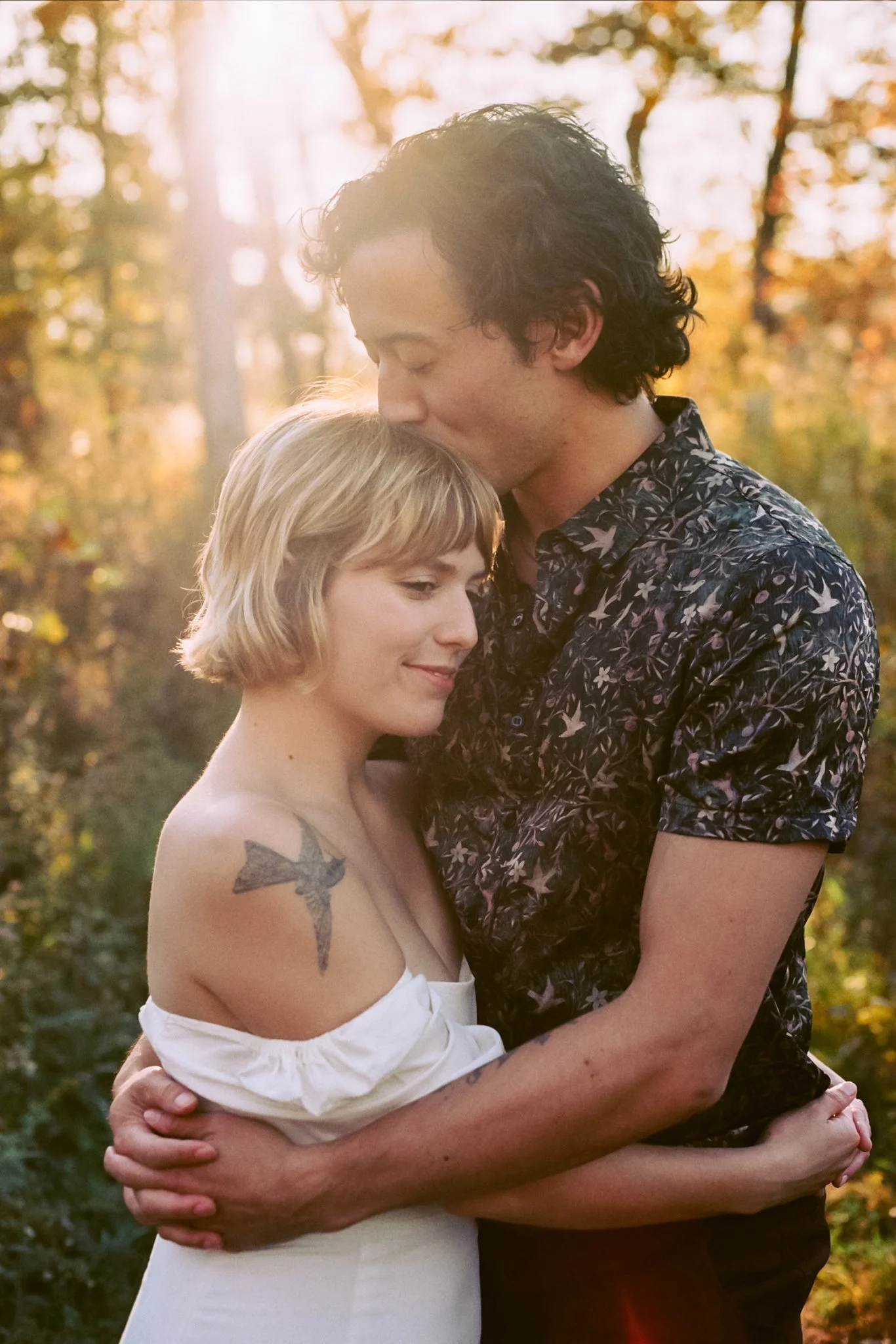 A couple embracing outdoors during sunset, with the man kissing the woman's forehead, both smiling gently, in a wooded area with golden trees.