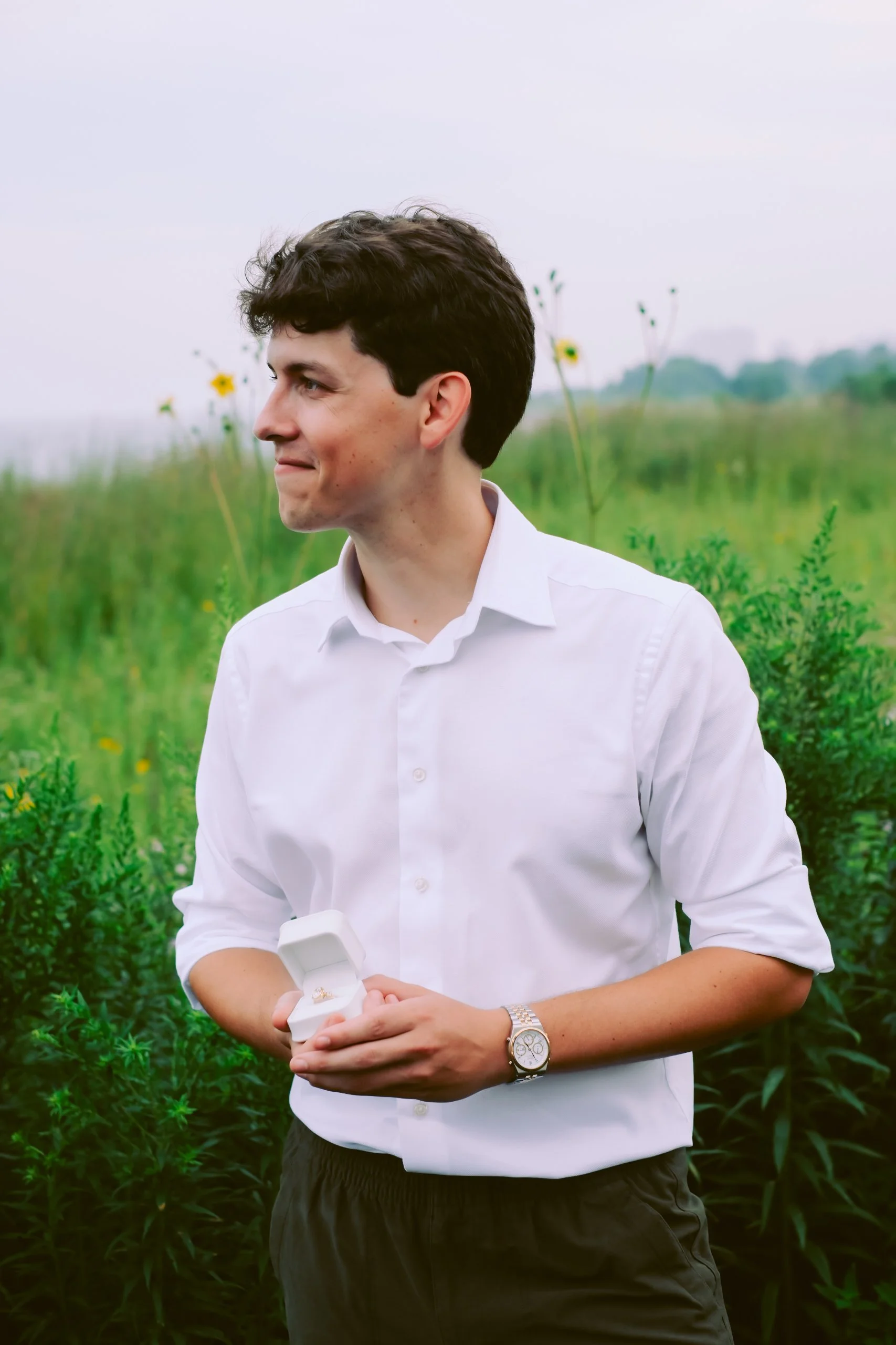 A man with dark hair wearing a white shirt and a watch is standing outdoors in a green field, holding an open jewelry box with a ring inside, and looking to his left with a slight smile.