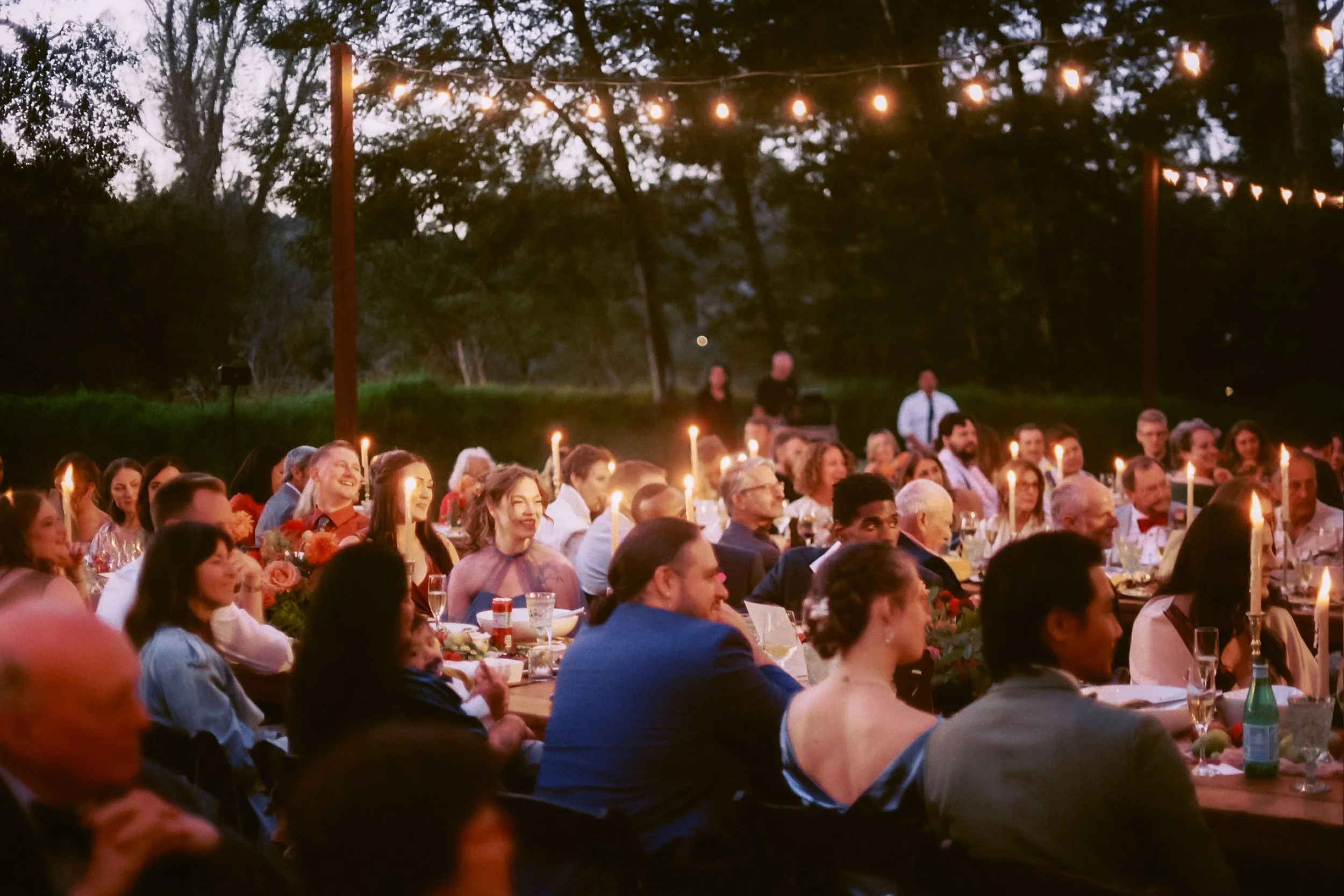 People gathered outdoors for a celebration or dinner, sitting at long tables with candles, under string lights at dusk or evening, surrounded by trees.