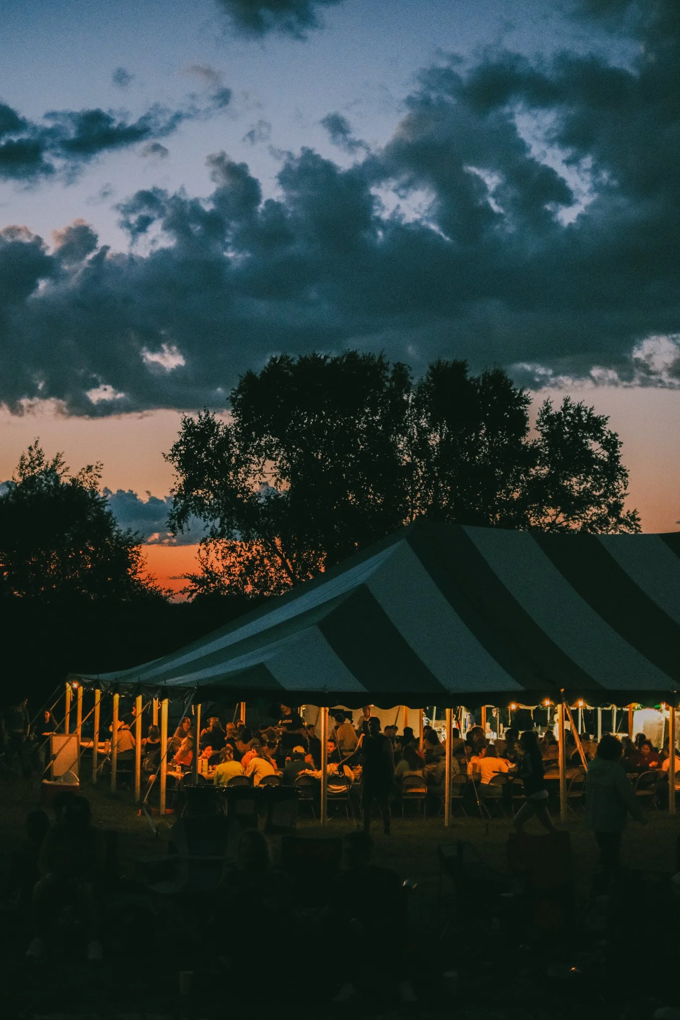 An outdoor evening gathering under a large striped tent with people seated at tables. The sky is dark with clouds, and the setting appears to be at sunset or dusk, with trees and a building partially visible in the background.